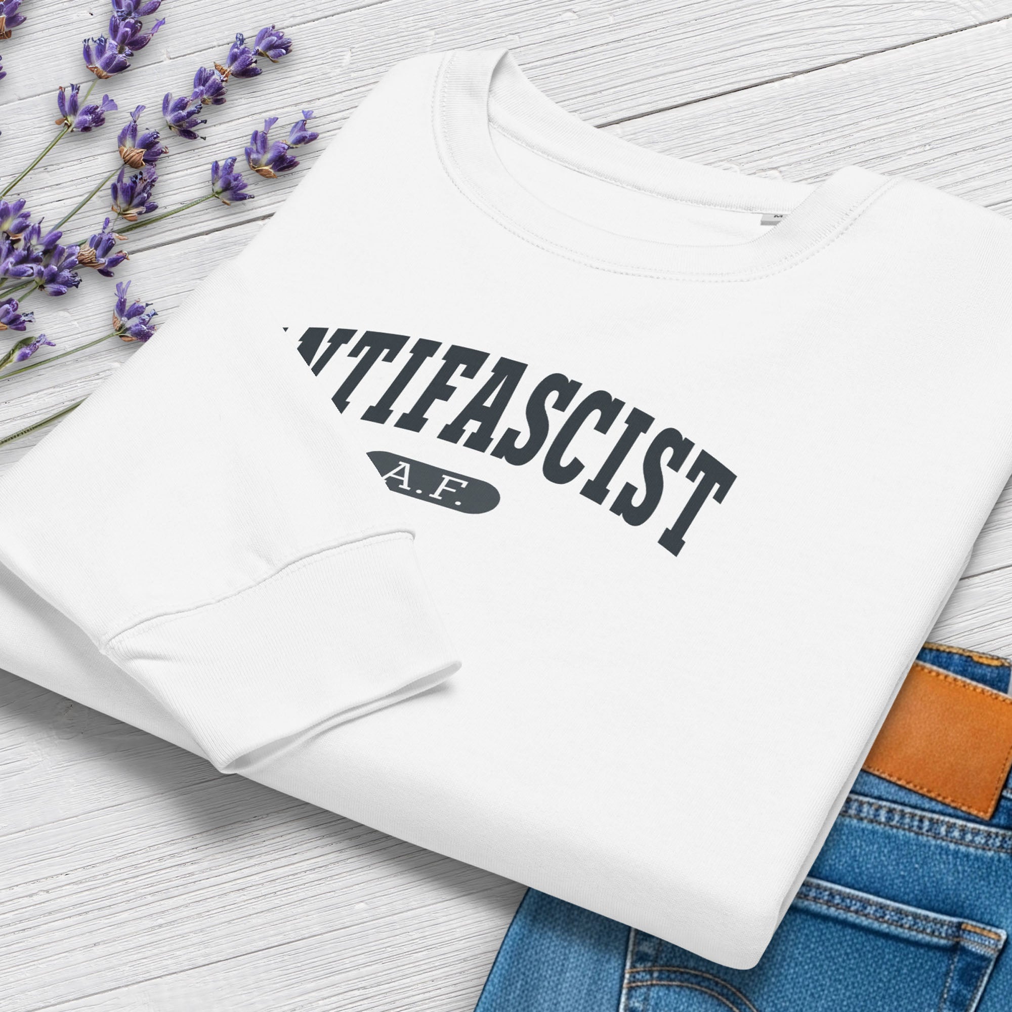 Folded white sweatshirt with dark grey text that reads “Antifascist A.F.” in a collegiate style. Shows closeup cuff detail, styled with greenery and jeans.