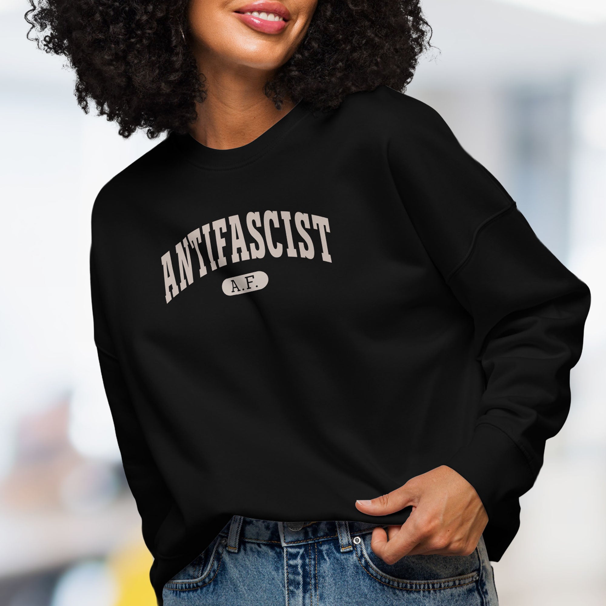 Front view of female model wearing black sweatshirt with greyish tan text that reads “Antifascist A.F.” in a collegiate style.
