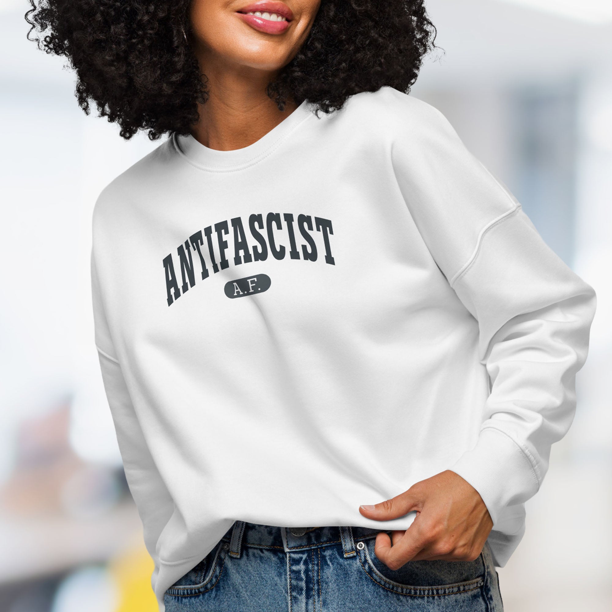 Front view of female model wearing white sweatshirt with dark grey text that reads “Antifascist A.F.” in a collegiate style.