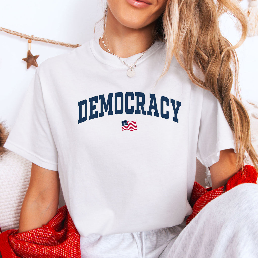 Female model wearing white t-shirt with the word “Democracy” in bold navy letters and a small American flag underneath, symbolizing U.S. pride in democracy over authoritarianism, tyranny, dictators.