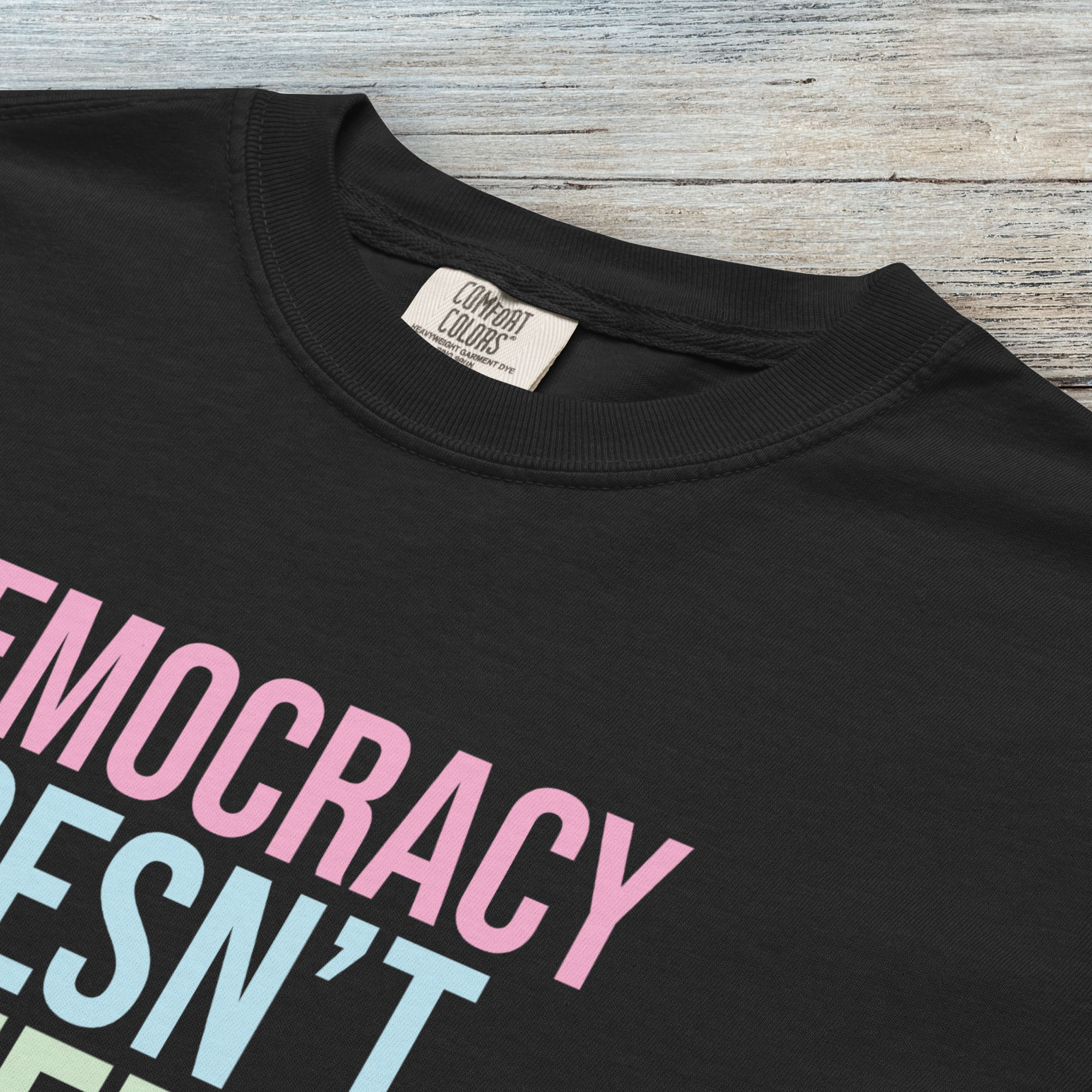 Closeup detail of black unisex political t-shirt that says “Democracy Doesn’t Defend Itself” in pink, blue, green, and yellow on a black Comfort Colors 1717 shirt.