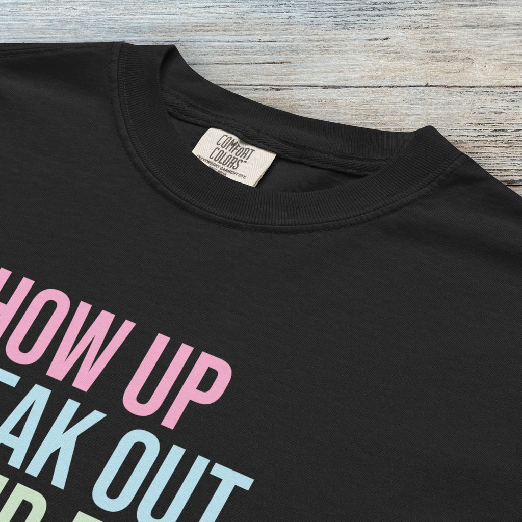 Closeup detail of black t-shirt with bright pink, blue and green bold text that says "show up, speak out, stand firm" and subtle grey script text below that reads "democracy doesn't defend itself."