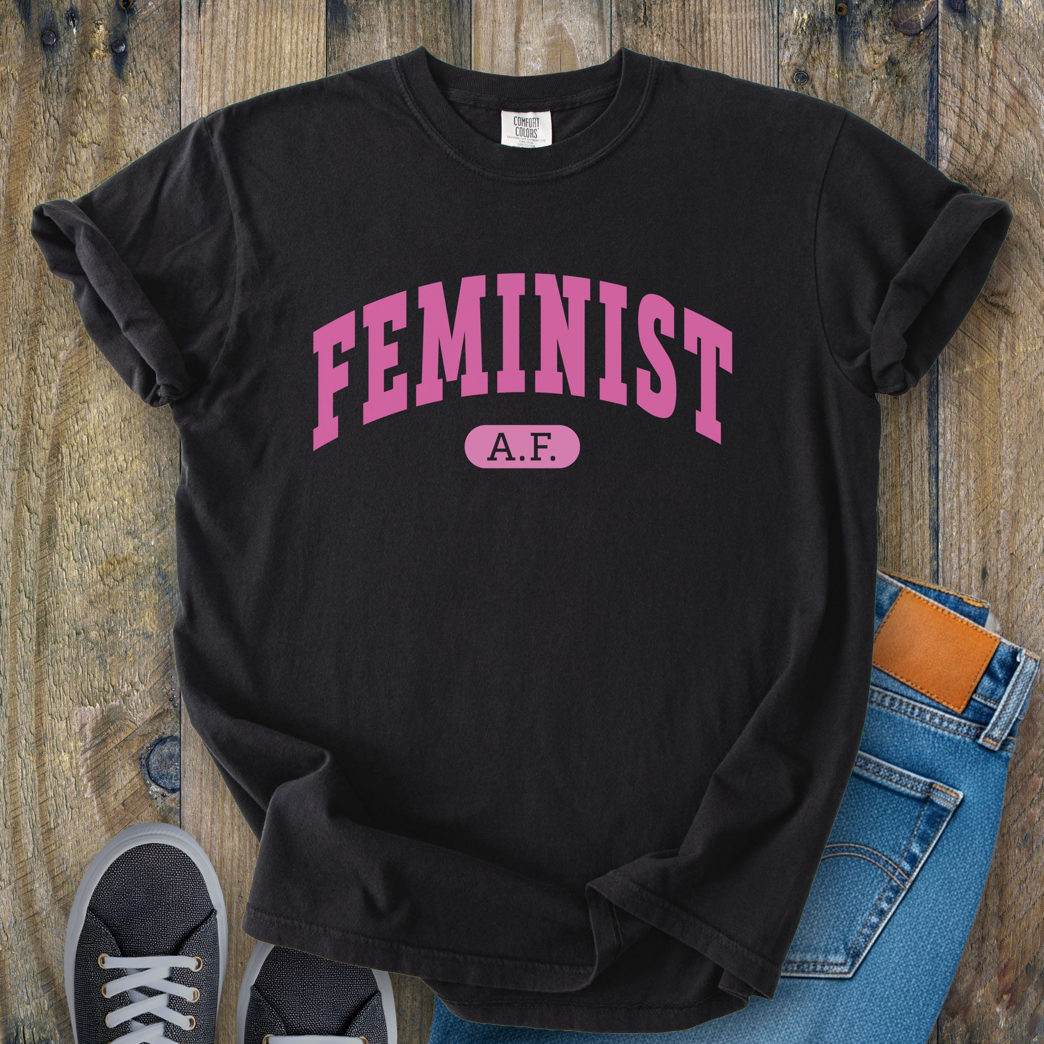 Black Comfort Colors tee with pink text that reads “Feminist A.F.” in a collegiate style.