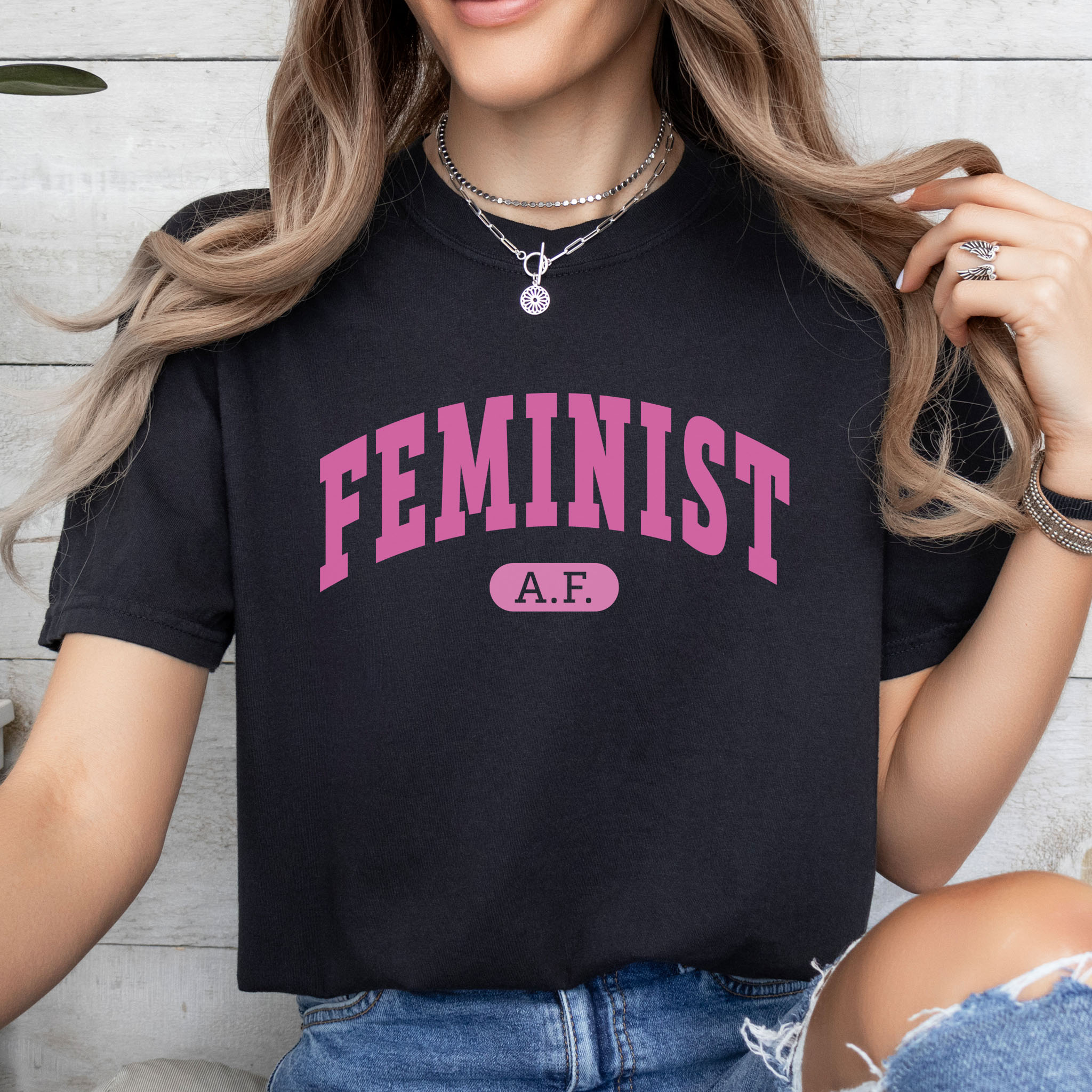 Female model wearing black Comfort Colors tee with pink text that reads “Feminist A.F.” in a collegiate style.