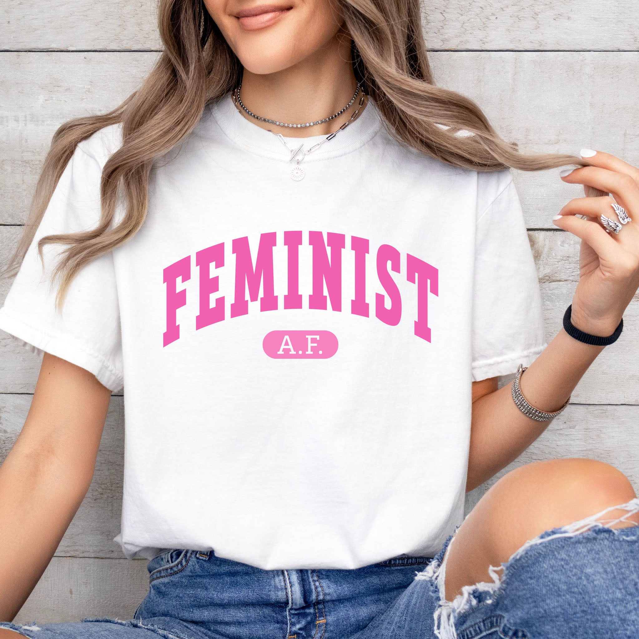 Female model wearing white Comfort Colors tee with pink text that reads “Feminist A.F.” in a collegiate style.