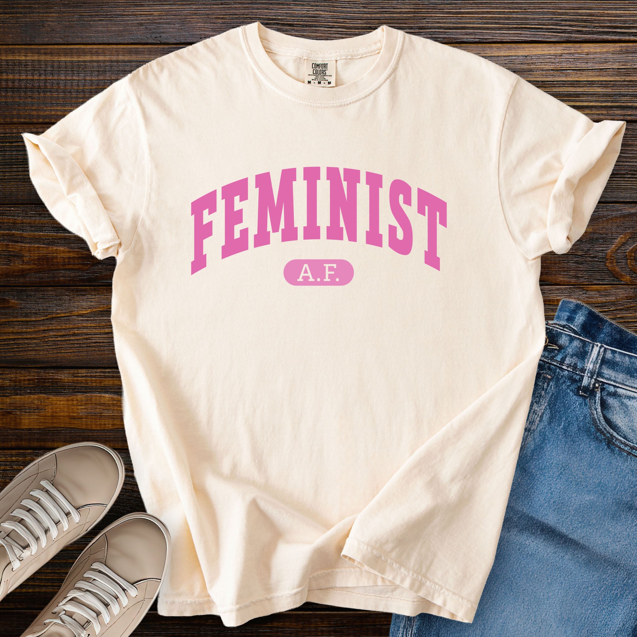 Ivory Comfort Colors tee with pink text that reads “Feminist A.F.” in a collegiate style.