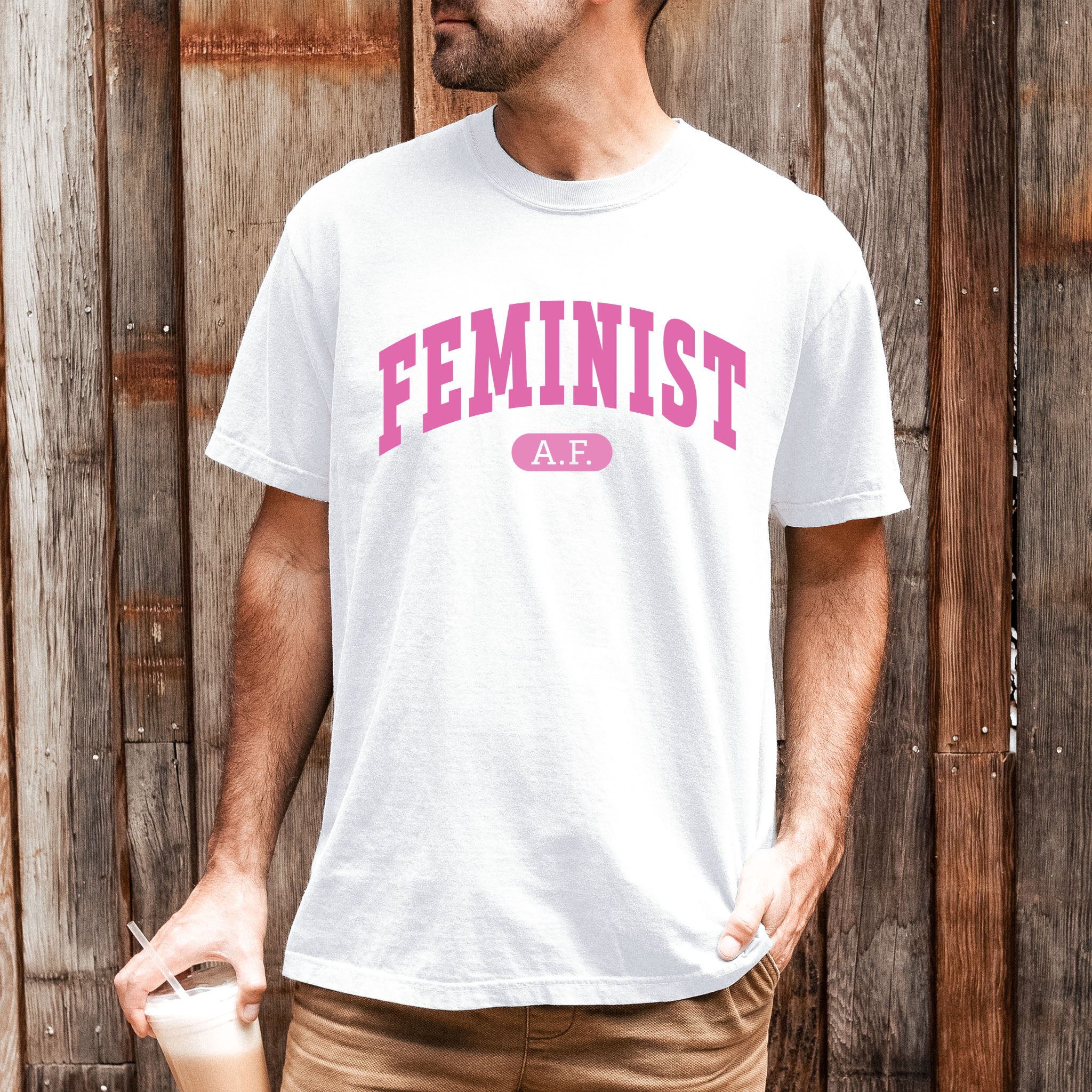 Male model wearing white Comfort Colors tee with pink text that reads “Feminist A.F.” in a collegiate style.