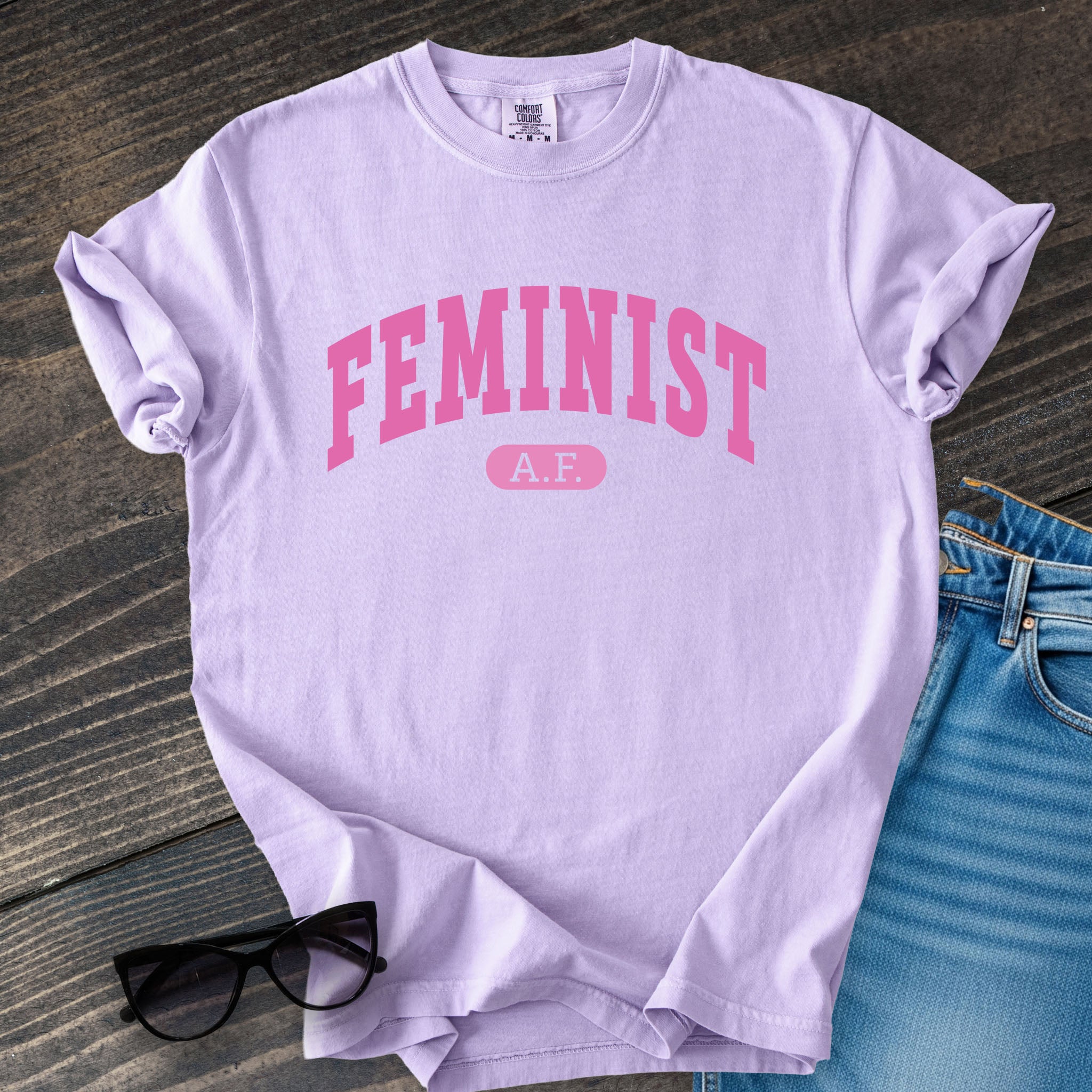 Orchid purple Comfort Colors tee with pink text that reads “Feminist A.F.” in a collegiate style.