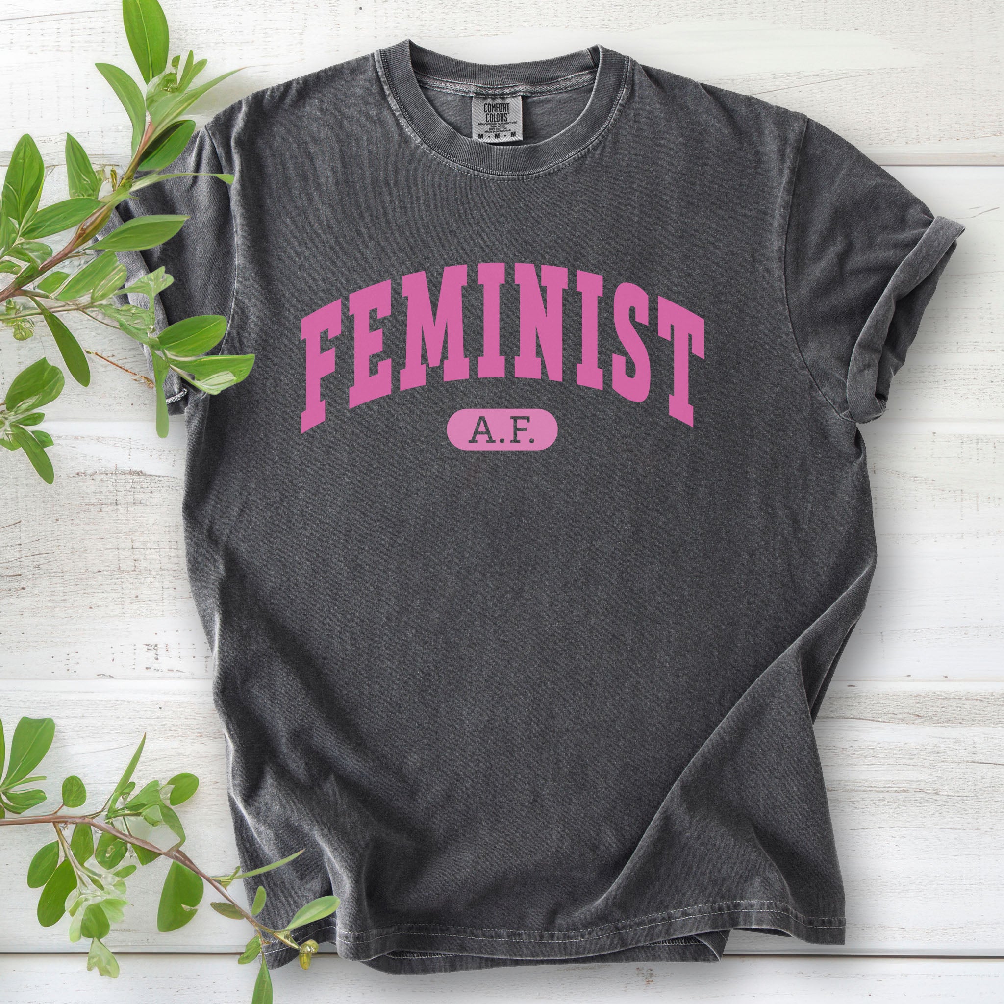 Pepper grey Comfort Colors tee with pink text that reads “Feminist A.F.” in a collegiate style.