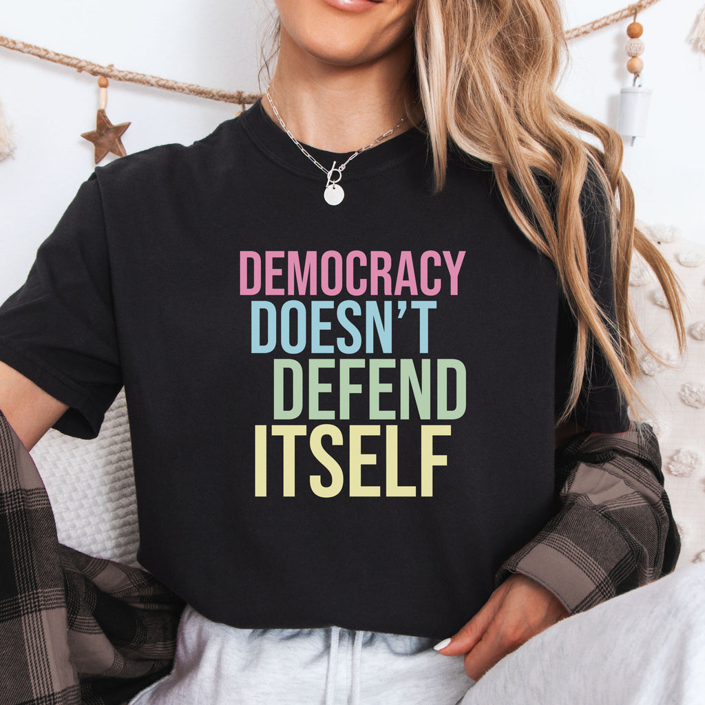 Female model wearing black unisex political t-shirt that says “Democracy Doesn’t Defend Itself” in pink, blue, green, and yellow on a black Comfort Colors 1717 shirt.