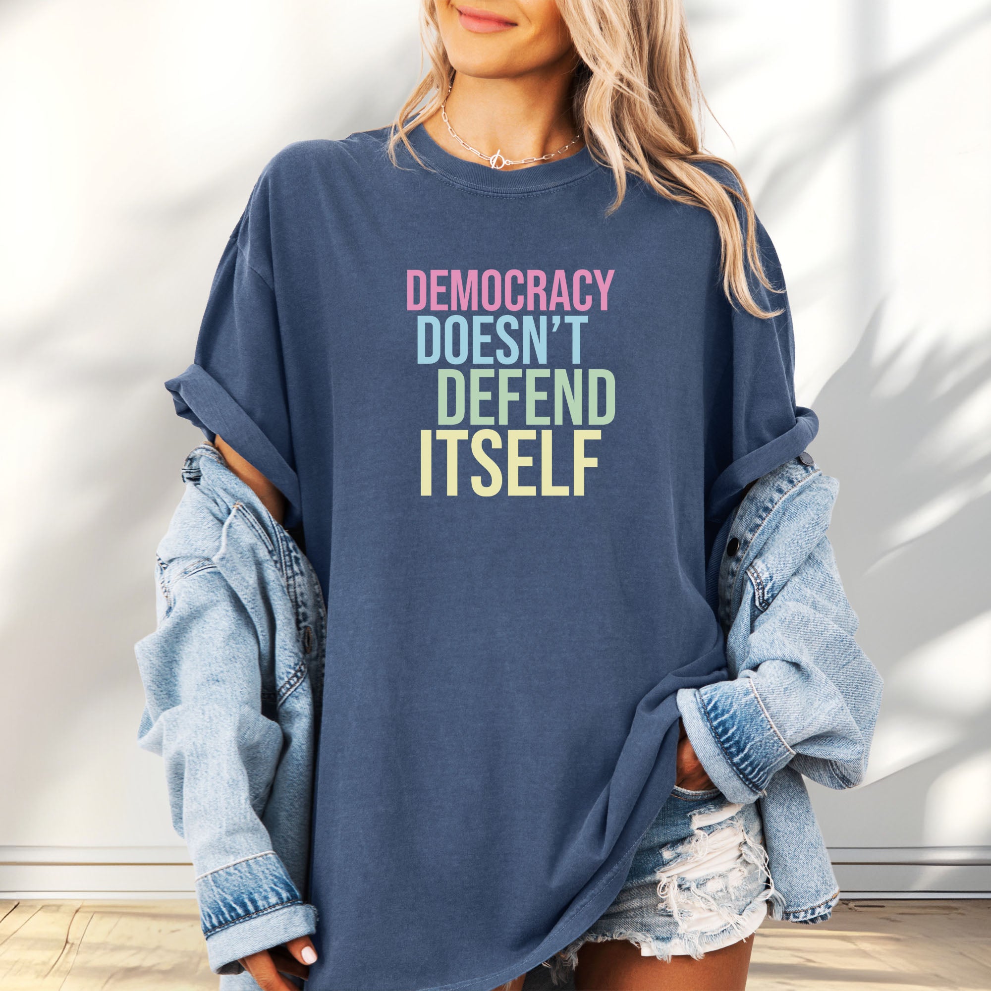 Female model wearing oversized denim unisex political t-shirt that says “Democracy Doesn’t Defend Itself” in pink, blue, green, and yellow on a black Comfort Colors tee.