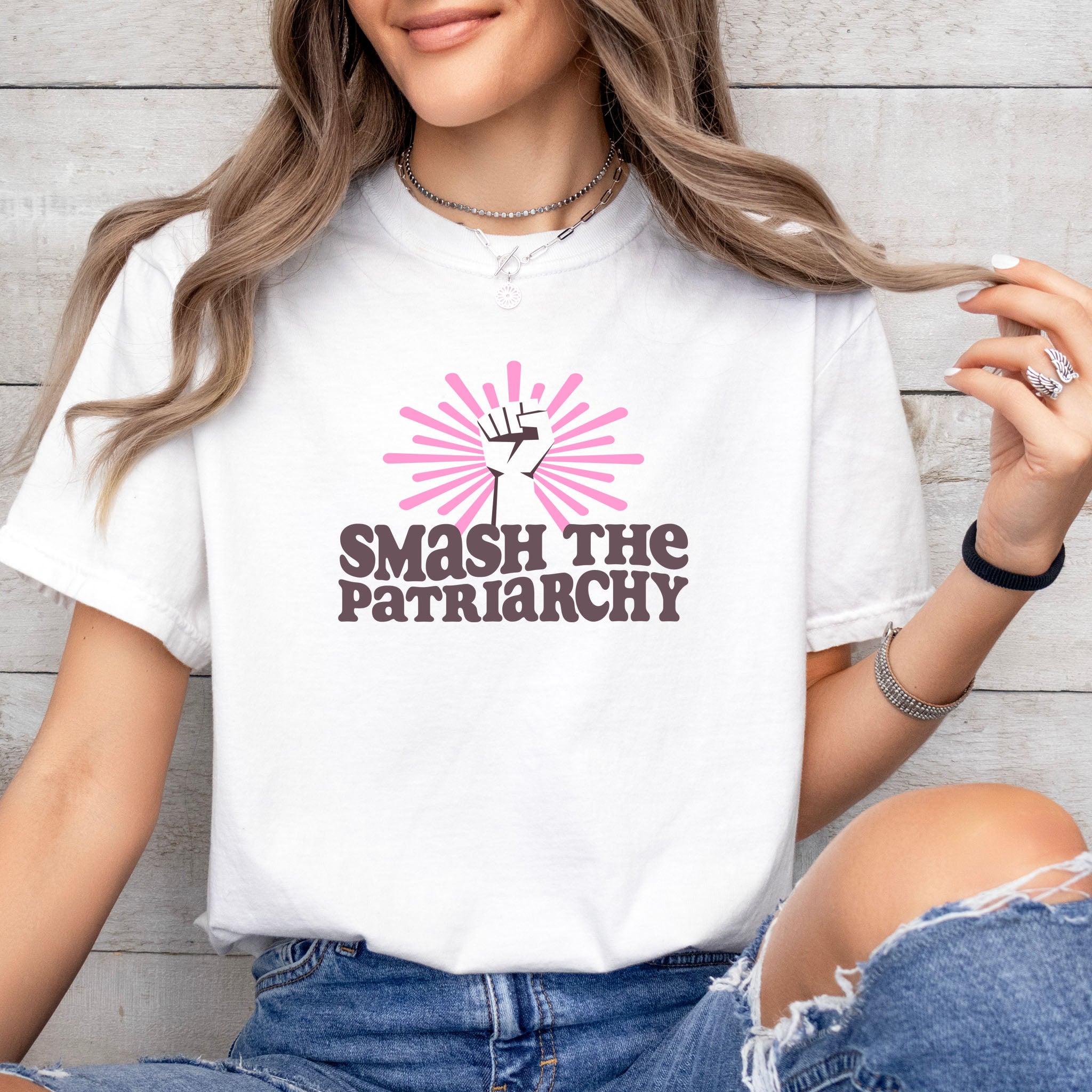 Female model wearing white t-shirt with a raised fist graphic and pink rays behind it, featuring the text “Smash the Patriarchy.”