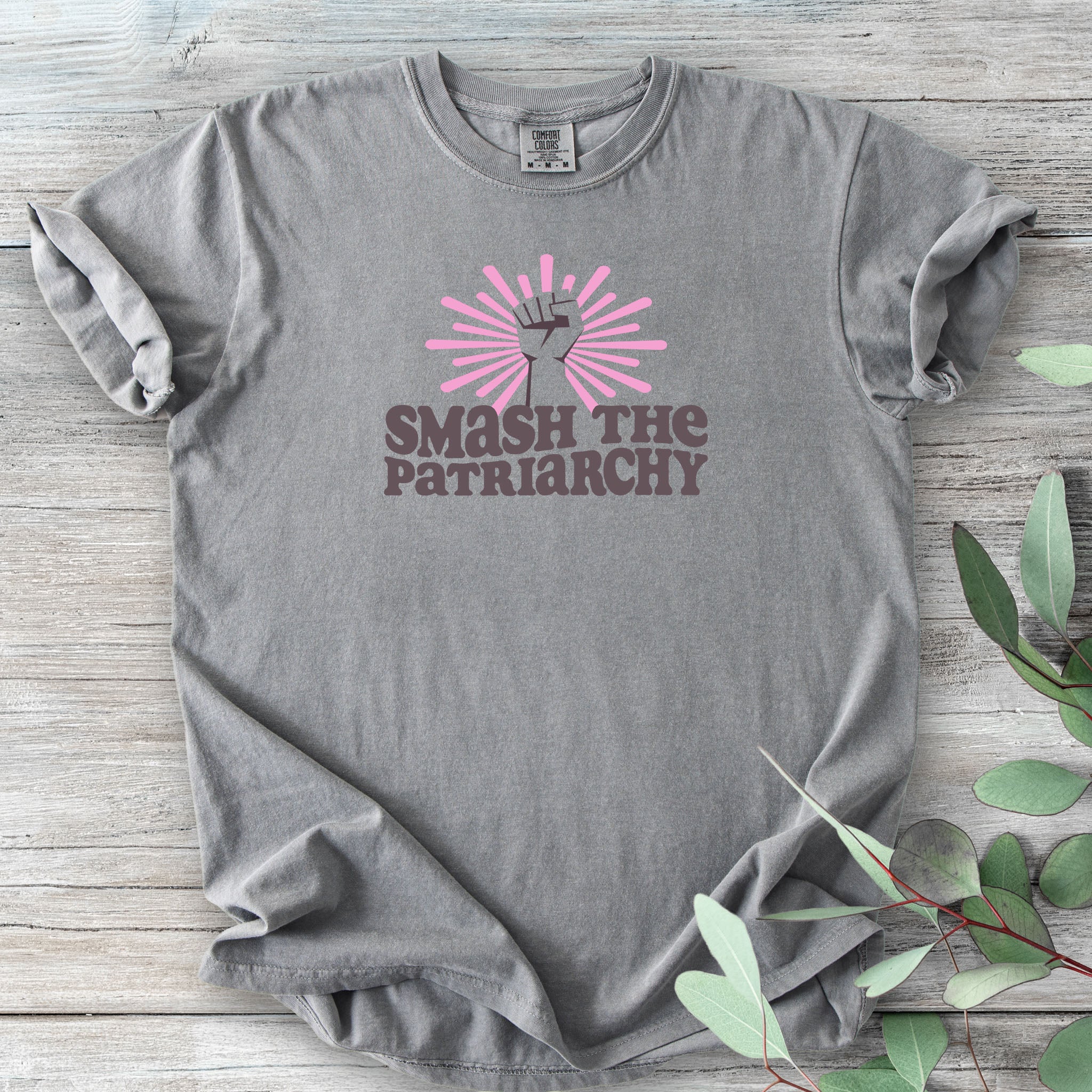 Grey t-shirt with a raised fist graphic and pink rays behind it, featuring the text “Smash the Patriarchy.”