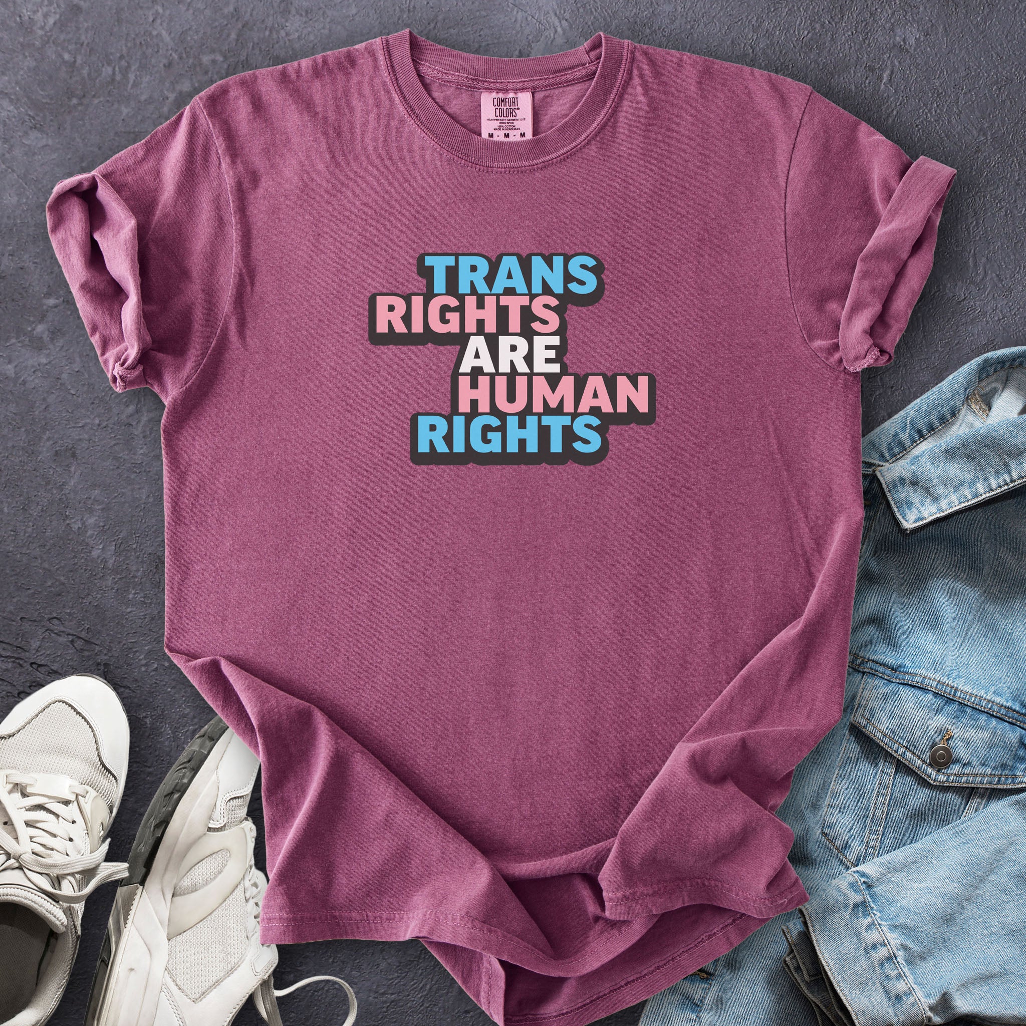 Berry color t-shirt with graphic that reads "trans rights are human rights" in the light blue, pink, and white colors of the trans flag, with a black shadow outlining the text.