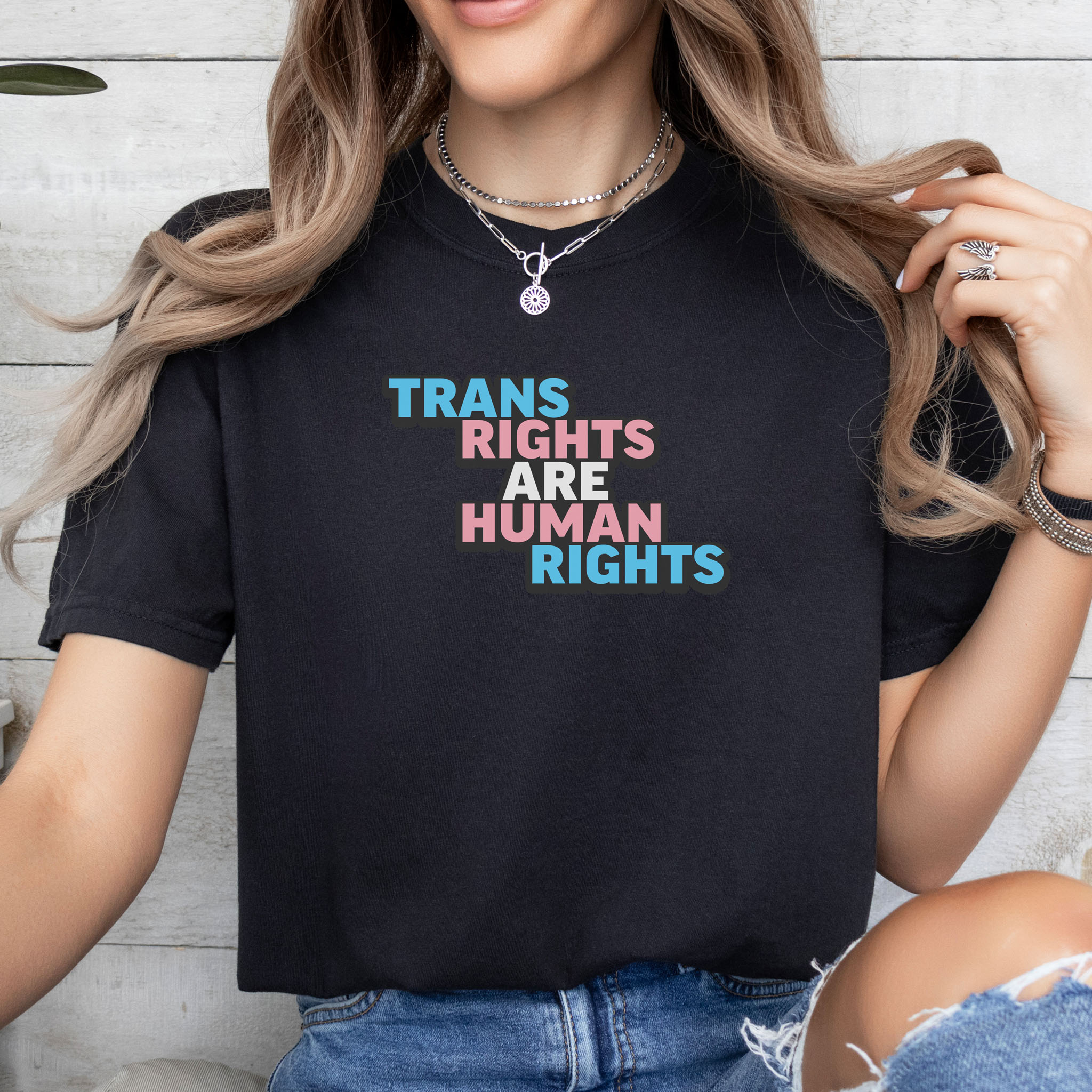 Female model wearing black t-shirt with graphic that reads "trans rights are human rights" in the light blue, pink, and white colors of the trans flag, with a black shadow outlining the text.