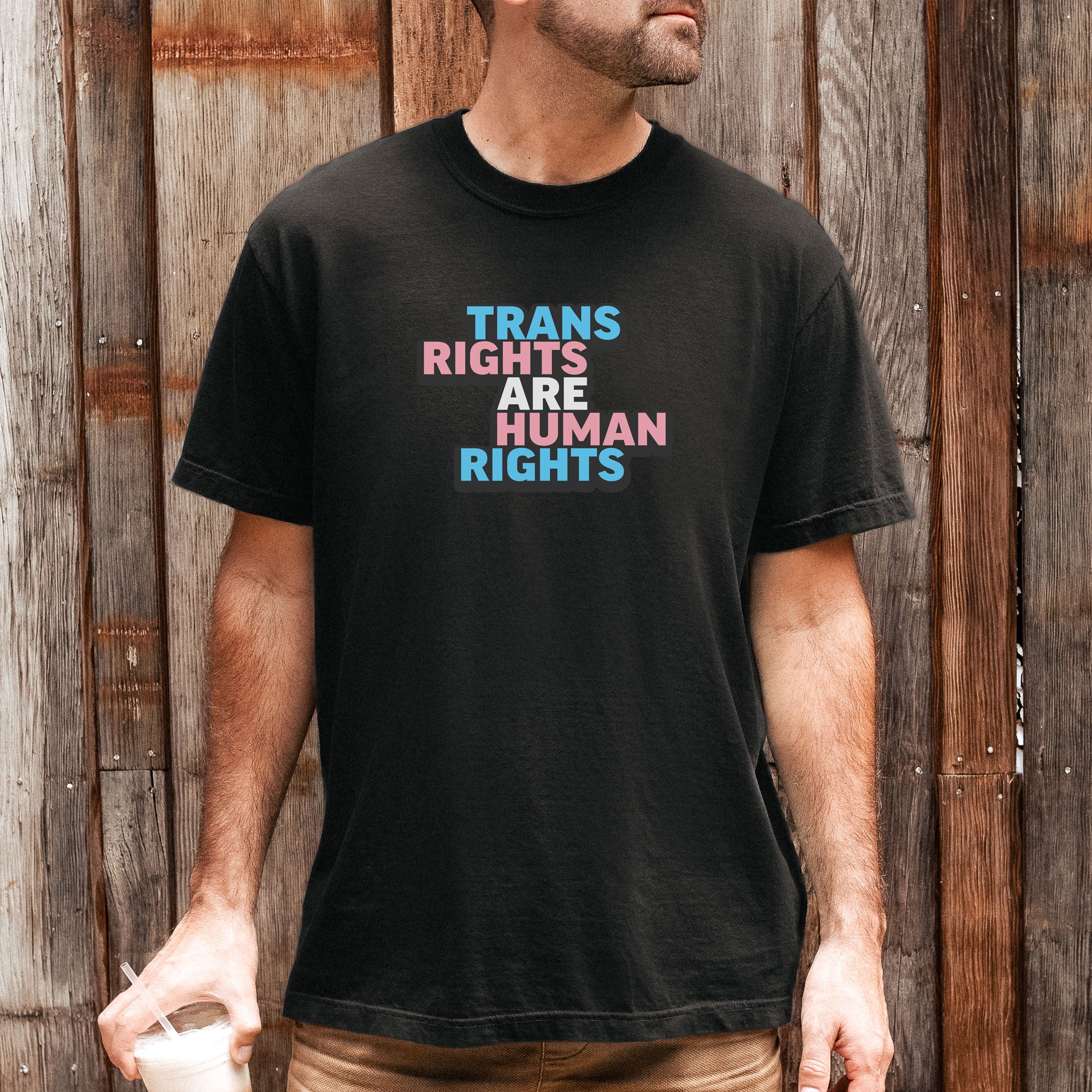 Male model wearing black t-shirt with graphic that reads "trans rights are human rights" in the light blue, pink, and white colors of the trans flag, with a black shadow outlining the text.