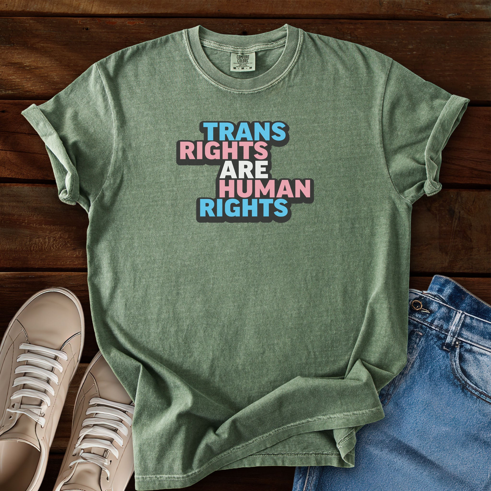 Moss green t-shirt with graphic that reads "trans rights are human rights" in the light blue, pink, and white colors of the trans flag, with a black shadow outlining the text.