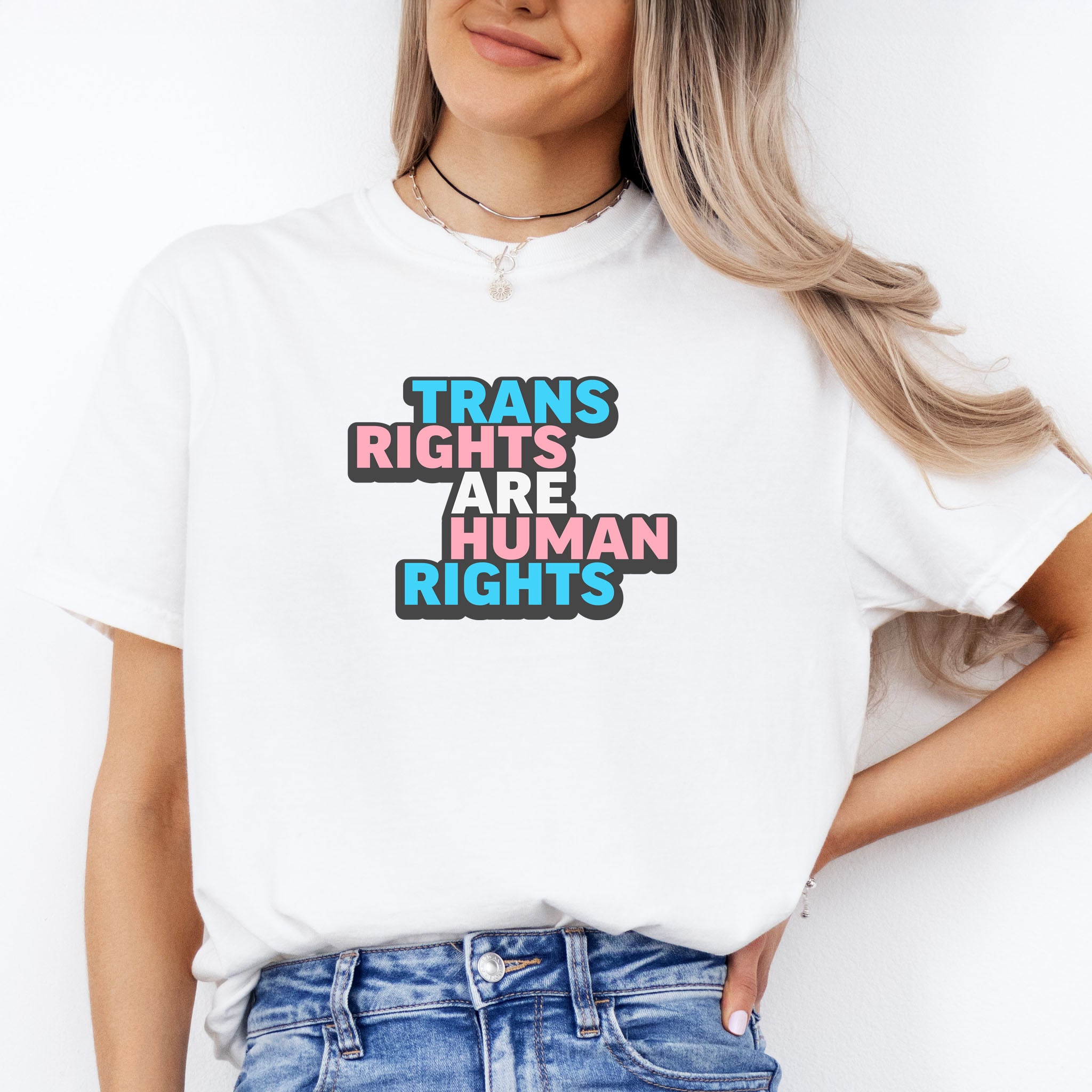 Female model wearing white color t-shirt with graphic that reads "trans rights are human rights" in the light blue, pink, and white colors of the trans flag, with a black shadow outlining the text.
