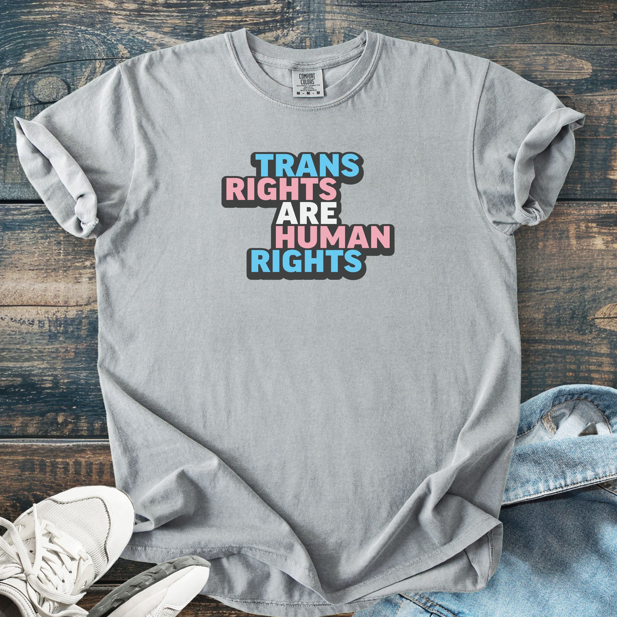 Gray color t-shirt with graphic that reads "trans rights are human rights" in the light blue, pink, and white colors of the trans flag, with a black shadow outlining the text.