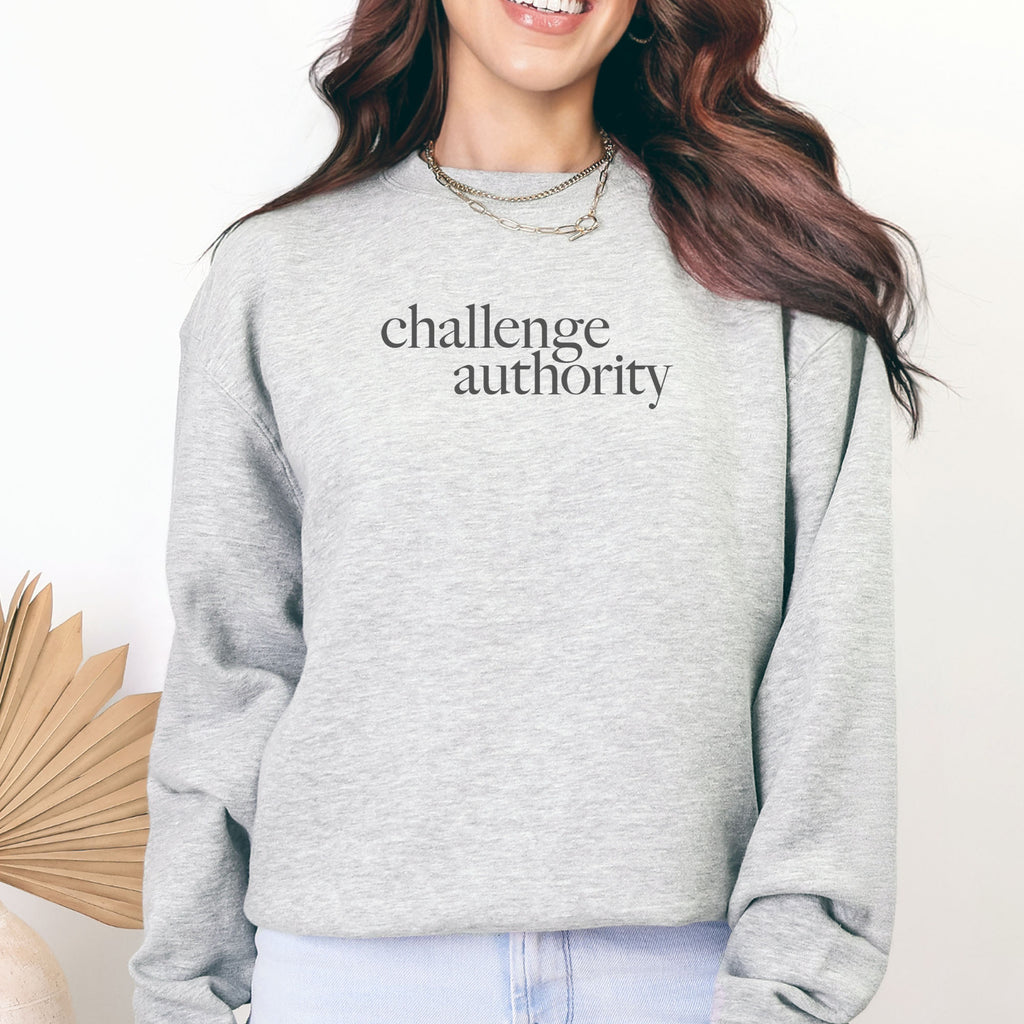 Female model wearing grey sweatshirt with minimalist serif text block reading “challenge authority."