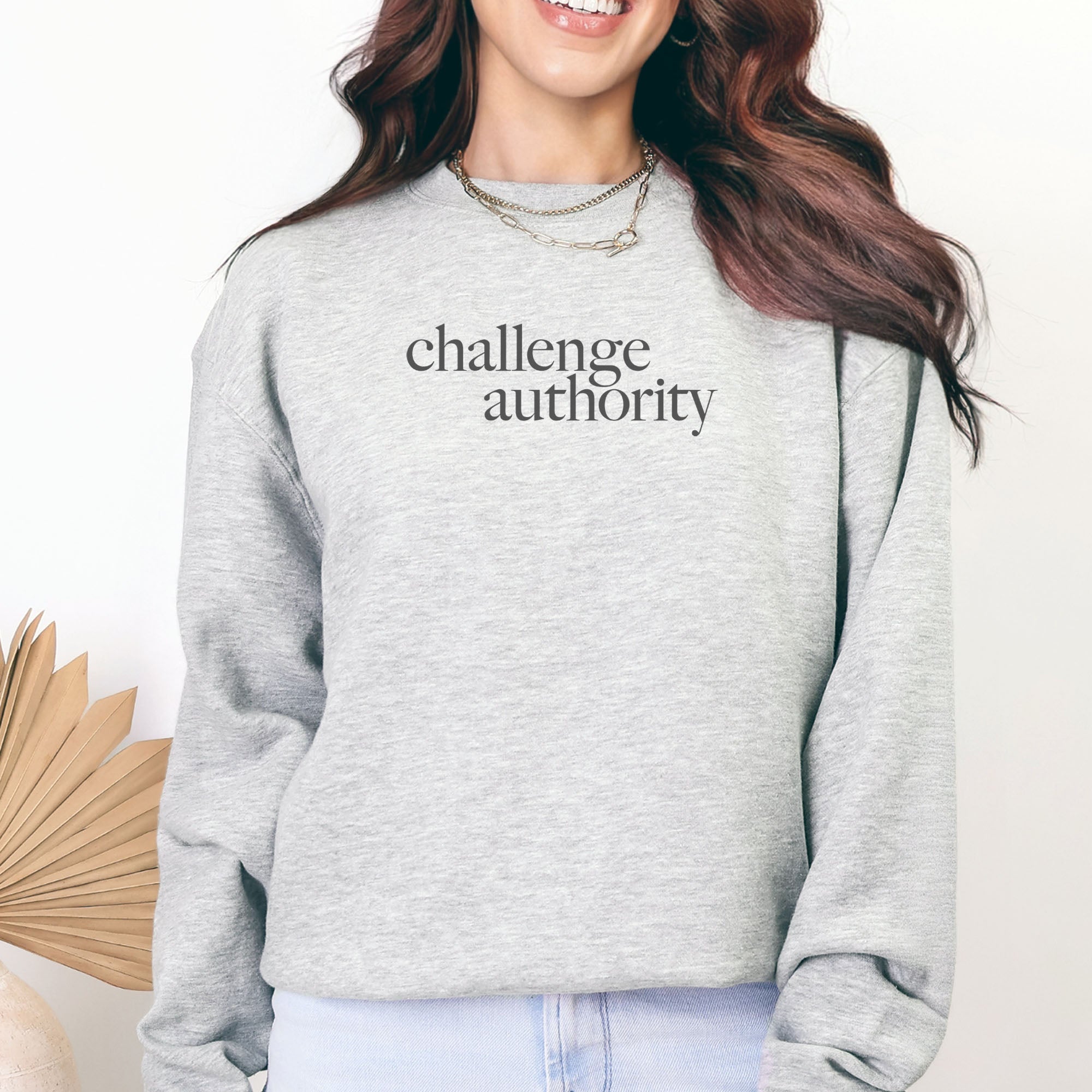 Female model wearing grey sweatshirt with minimalist serif text block reading “challenge authority."