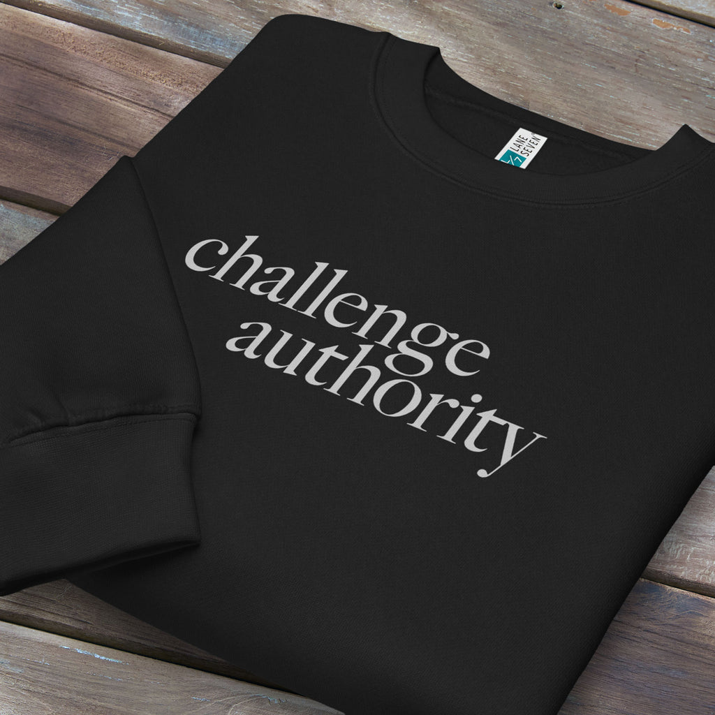 Folded black sweatshirt with minimalist serif text block reading “challenge authority."