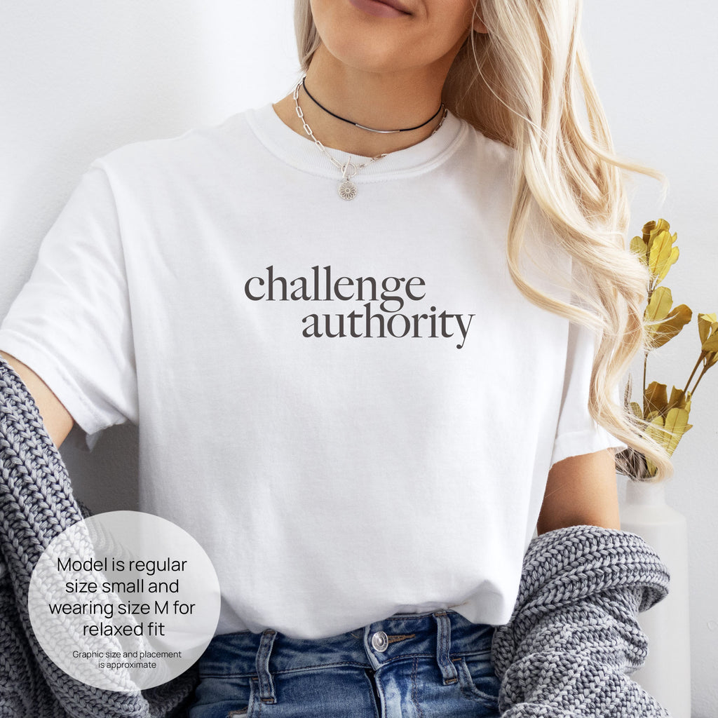 Female model wearing white t-shirt with minimalist serif text block reading “challenge authority." Includes size and fit information for the model who is regular size small, and wearing a size M shirt for a relaxed fit.
