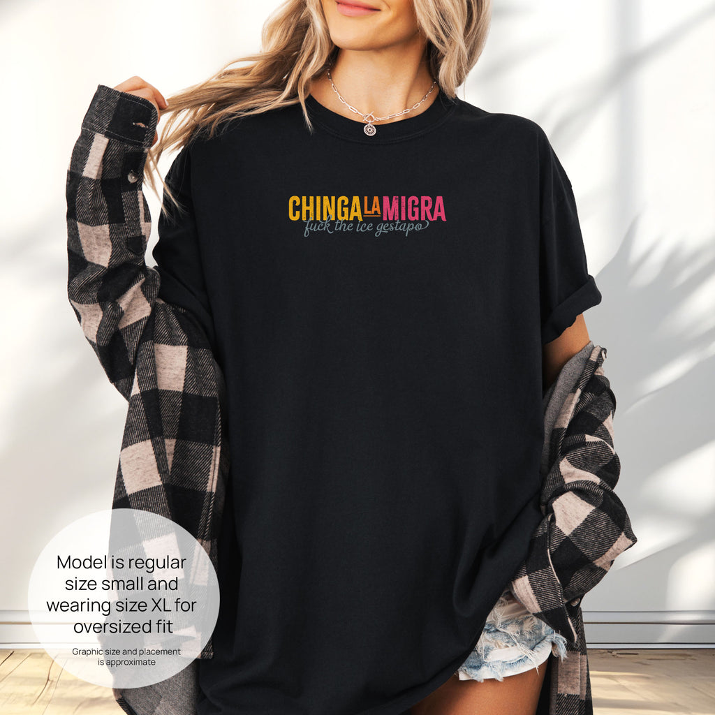 Female mode wearing black t-shirt with brightly colored text that reads "chinga la migra", with subtle script text below that says "fuck the ice gestapo." Includes size and fit information for the model who is regular size small, and wearing a size XL shirt for an oversized fit.
