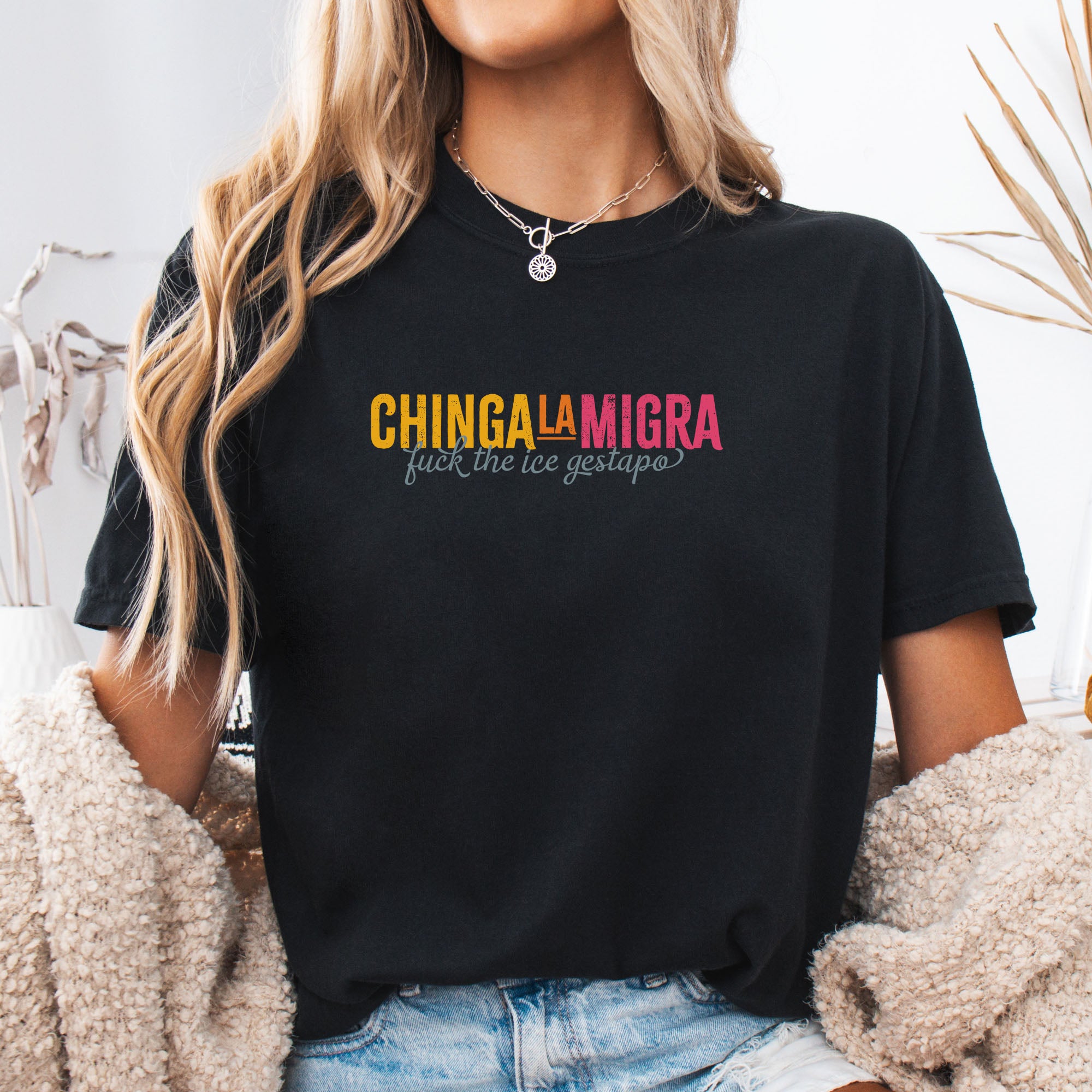 Female mode wearing black t-shirt with brightly colored text that reads "chinga la migra", with subtle script text below that says "fuck the ice gestapo."