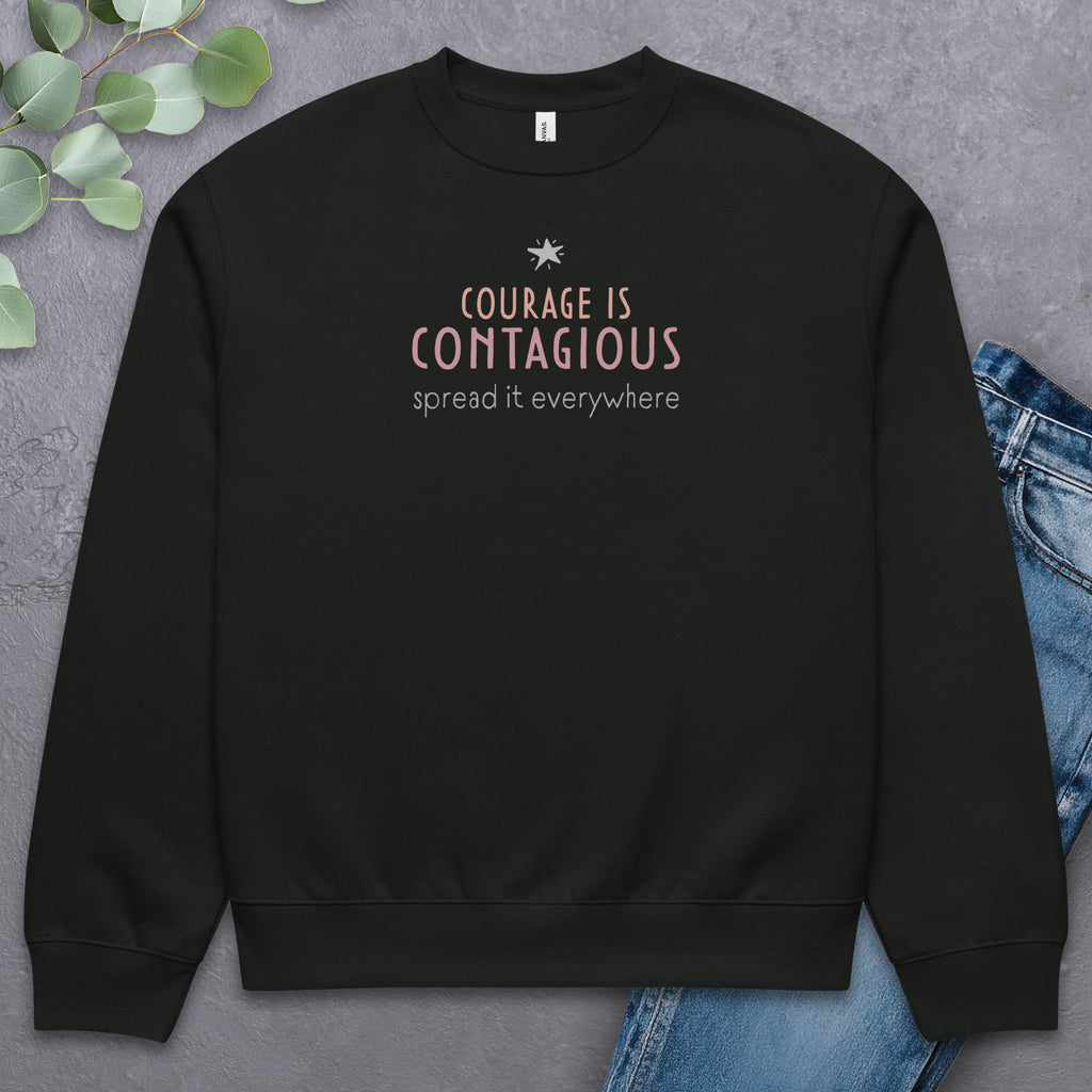 Black crewneck sweatshirt with minimalist stylized text that reads "courage is contagious – spread it everywhere" laying flat with a pair of jeans.