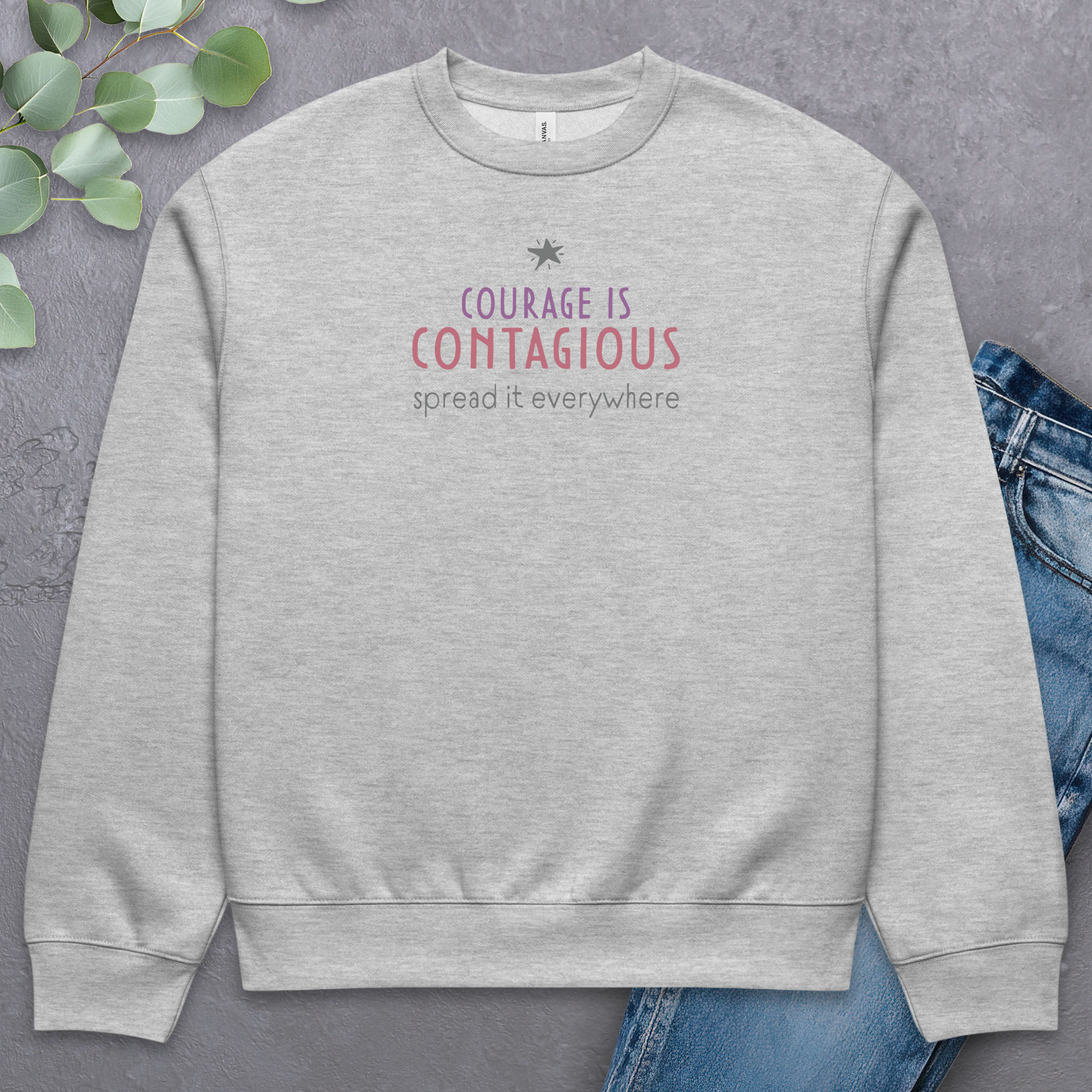 Athletic heather grey crewneck sweatshirt with minimalist stylized text that reads "courage is contagious – spread it everywhere" laying flat with a pair of jeans.