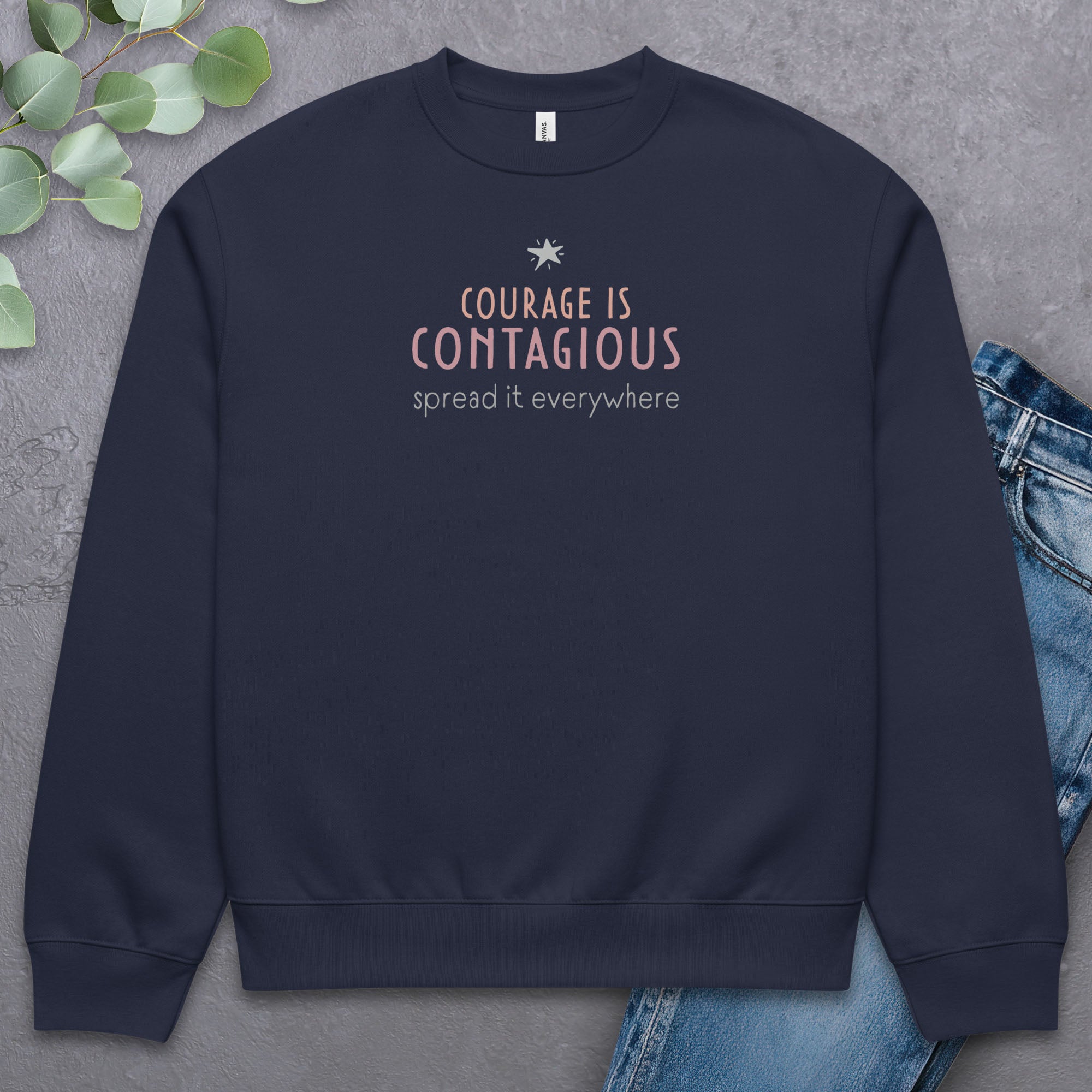 Navy crewneck sweatshirt with minimalist stylized text that reads "courage is contagious – spread it everywhere" laying flat with a pair of jeans.