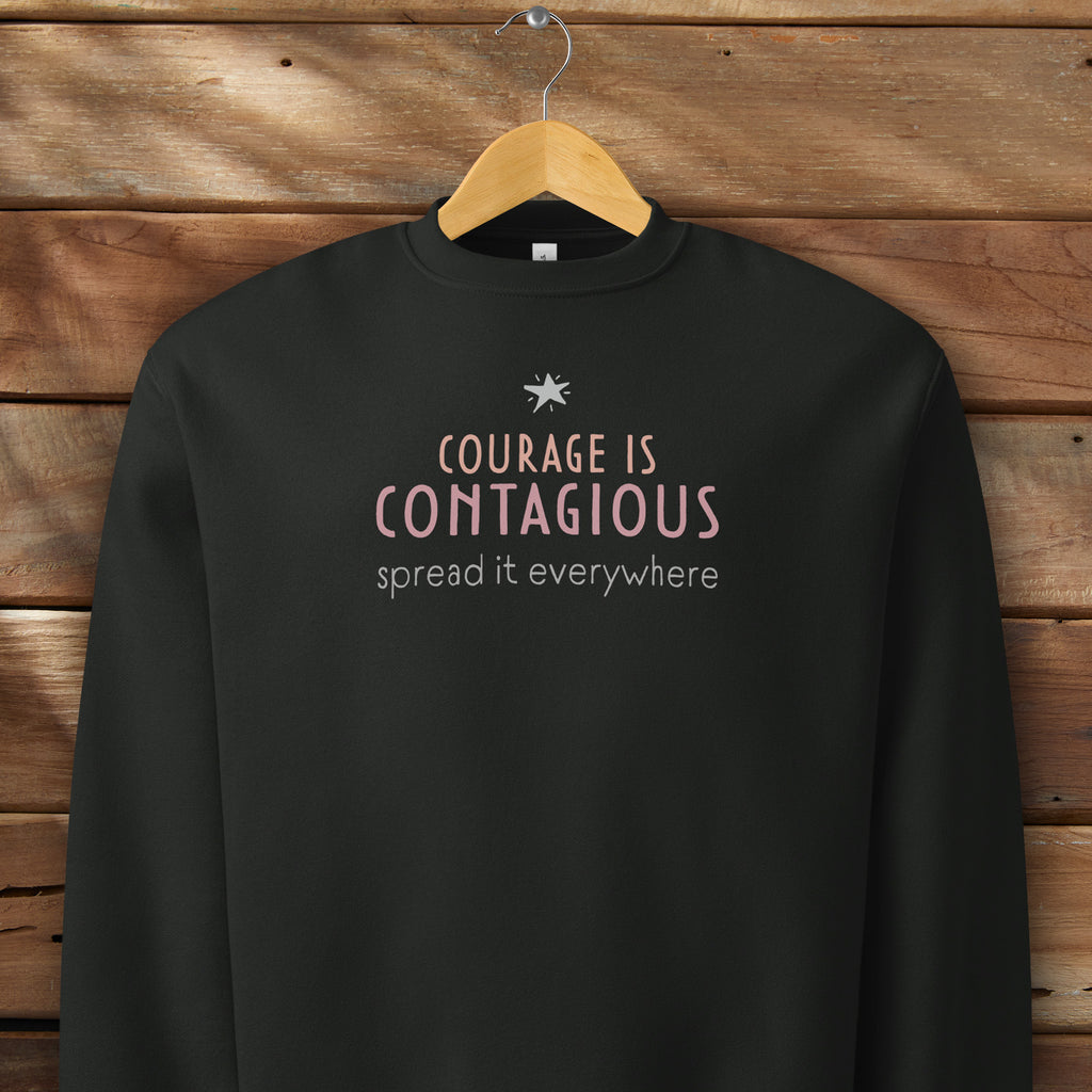 Black crewneck sweatshirt with minimalist stylized text that reads "courage is contagious – spread it everywhere" hanging on a hanger.