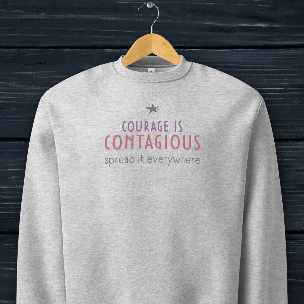 Athletic heather grey crewneck sweatshirt with minimalist stylized text that reads "courage is contagious – spread it everywhere" hanging on a hanger.