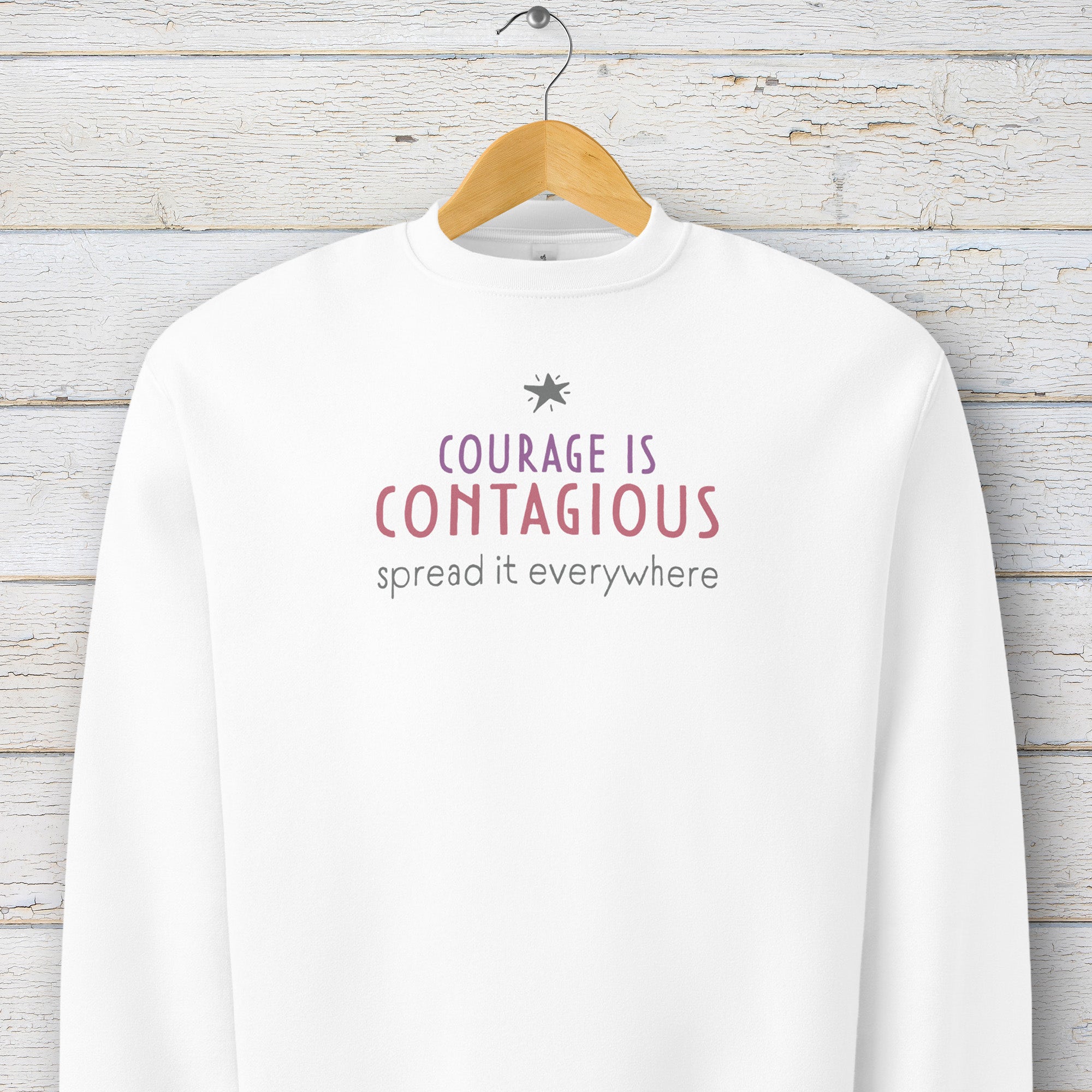 White crewneck sweatshirt with minimalist stylized text that reads "courage is contagious – spread it everywhere" hanging on a hanger.