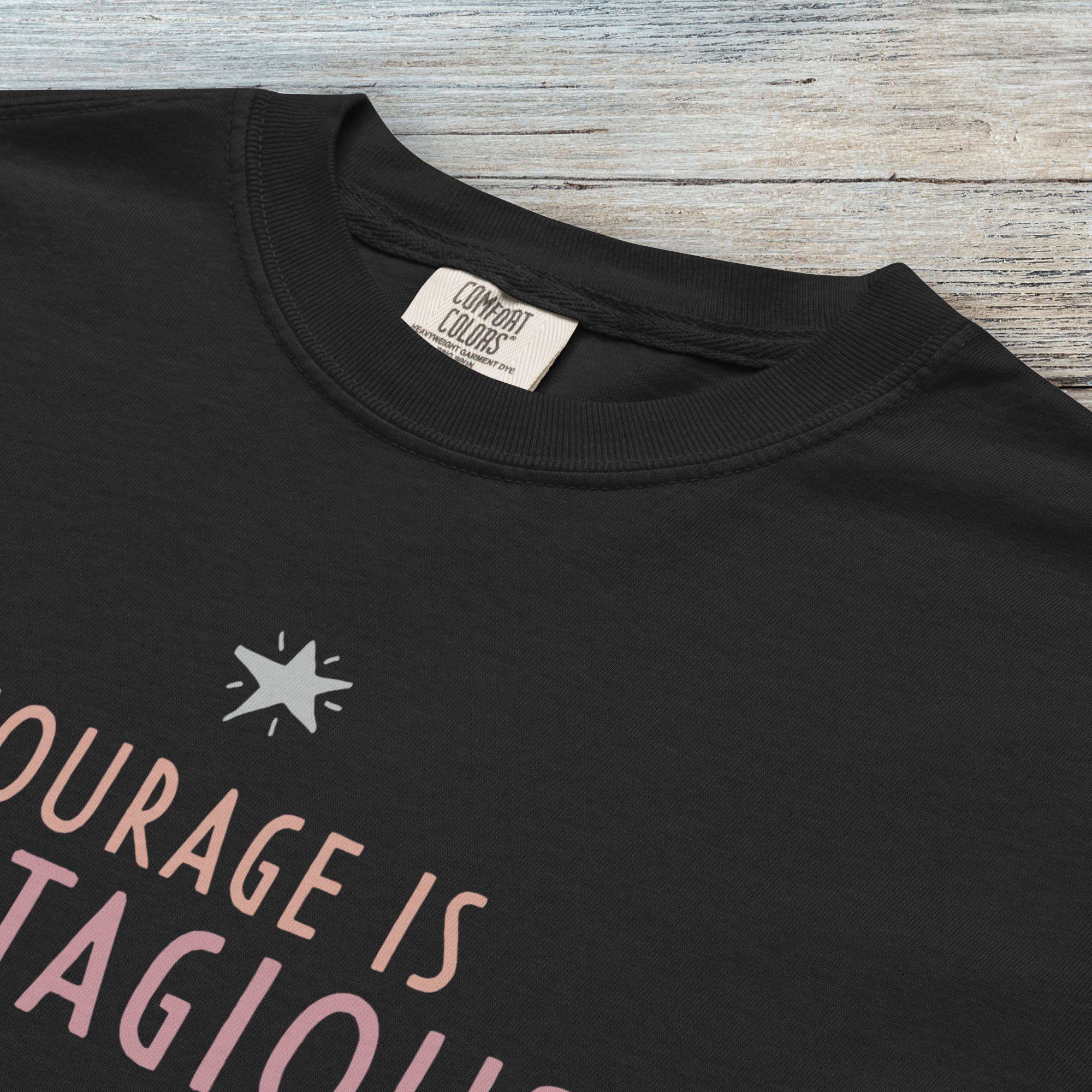 Closeup detail of black t-shirt with the words “Courage Is Contagious – Spread It Everywhere” printed in soft pink, coral, and gray.