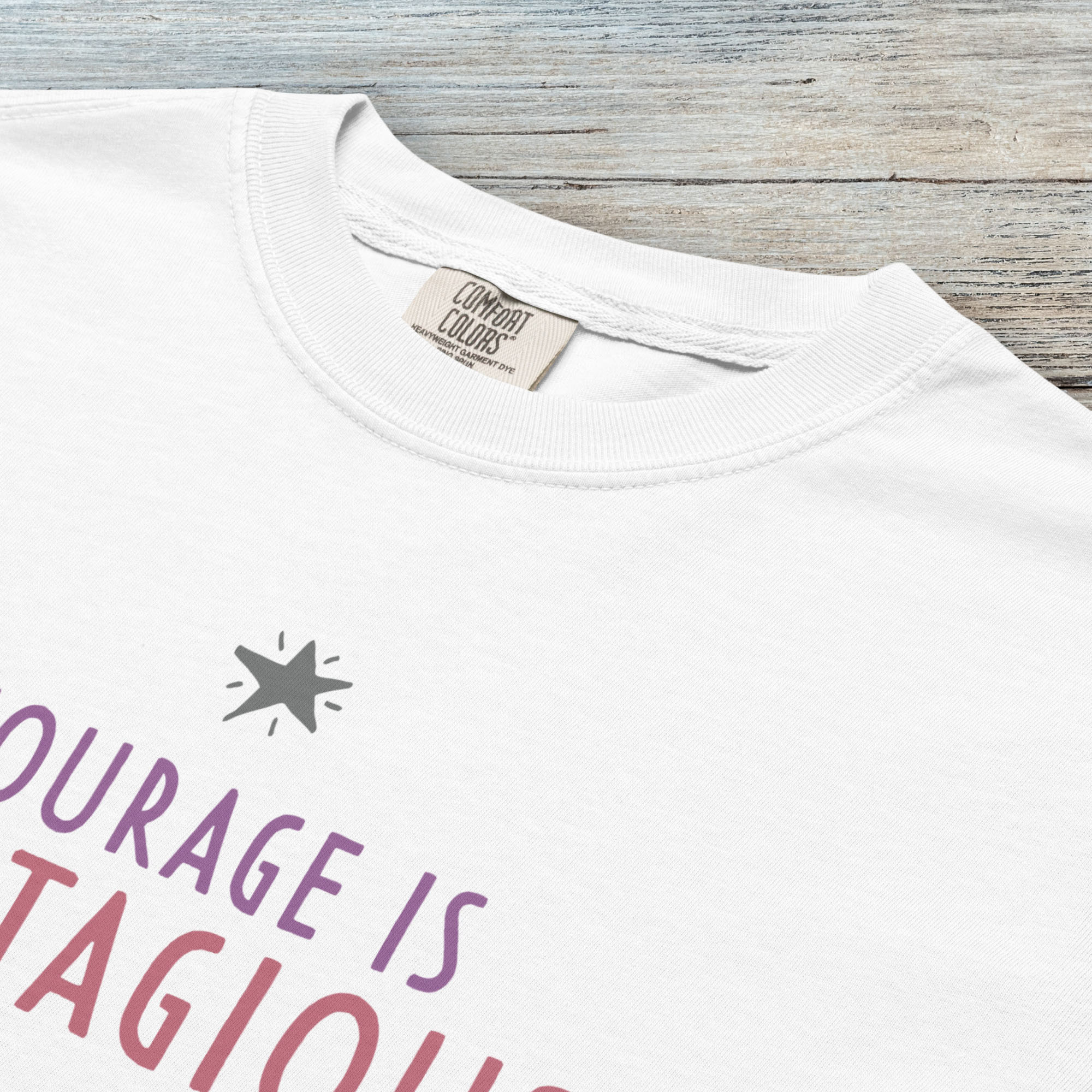 Closeup detail of white t-shirt with the words “Courage Is Contagious – Spread It Everywhere” printed in soft purple, red, and gray.