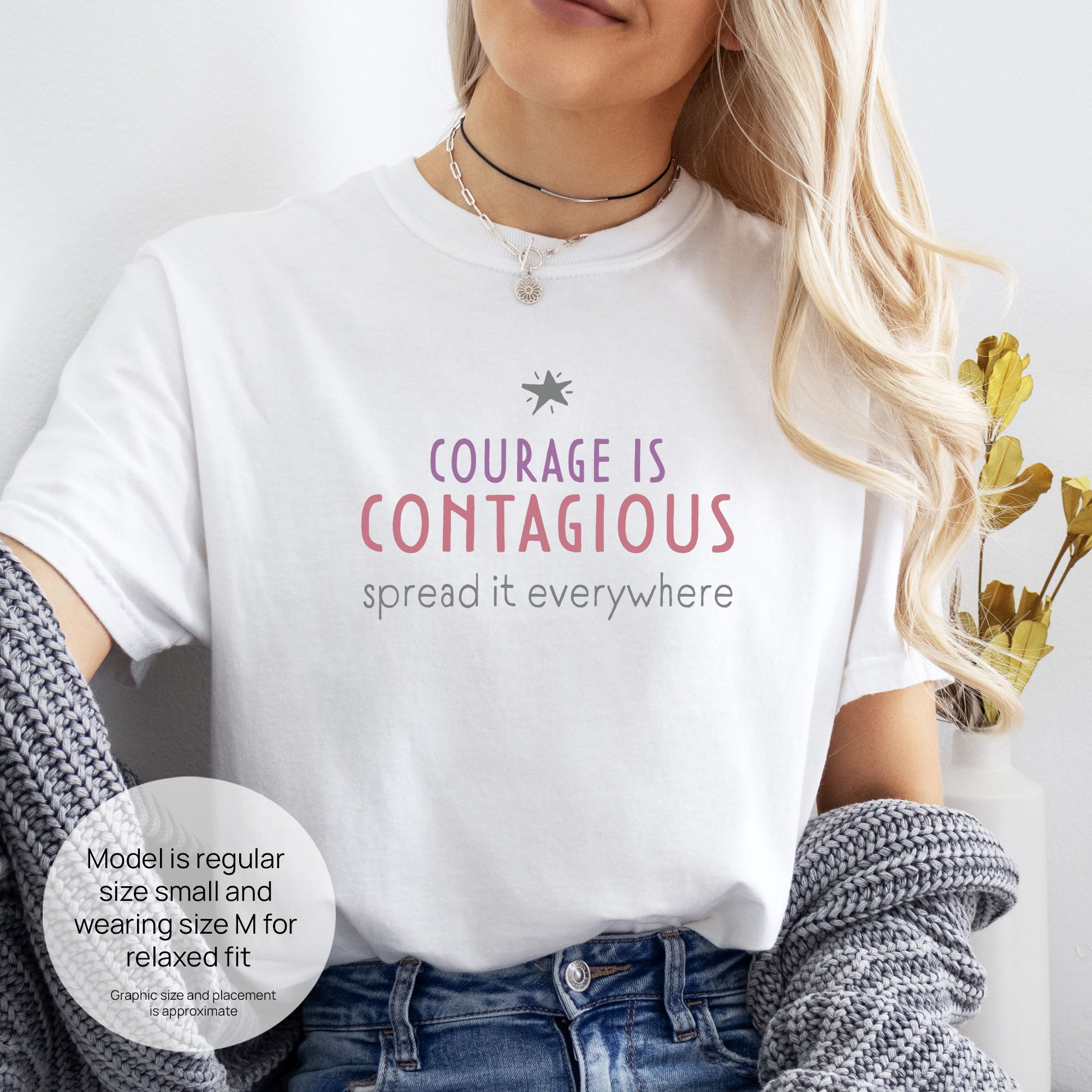Female model wearing white t-shirt with the words “Courage Is Contagious – Spread It Everywhere”. Includes size and fit information for the model who is regular size small, and wearing a size M shirt for a relaxed fit.
