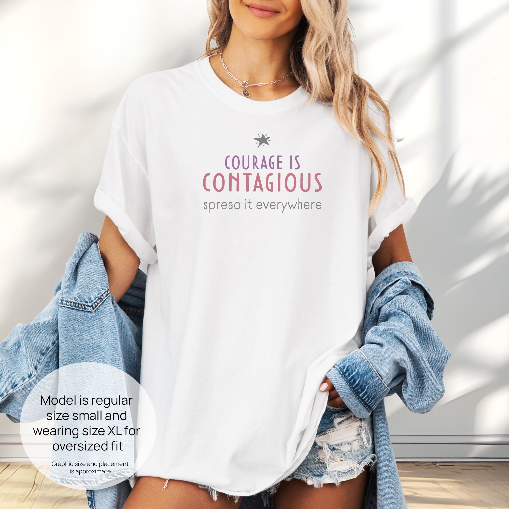 Female model wearing white t-shirt with the words “Courage Is Contagious – Spread It Everywhere”. Includes size and fit information for the model who is regular size small, and wearing a size XL shirt for an oversized fit.
