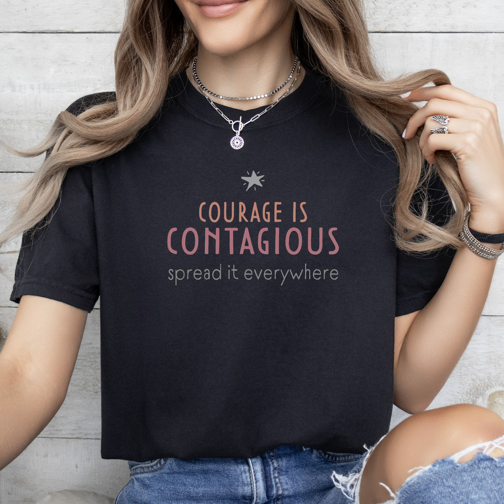Female model wearing black t-shirt with the words “Courage Is Contagious – Spread It Everywhere” printed in soft pink, coral, and gray.