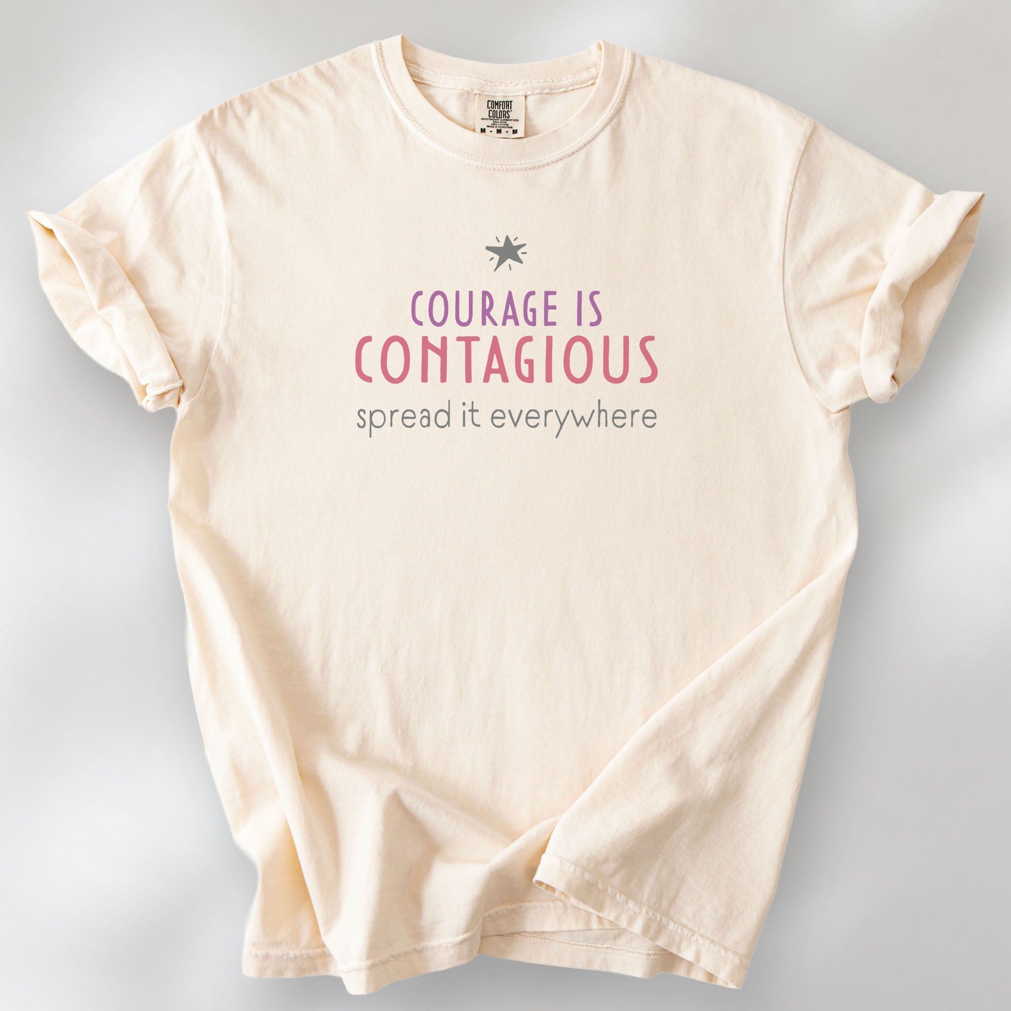 Ivory t-shirt with the words “Courage Is Contagious – Spread It Everywhere” printed in soft purple, red, and gray.