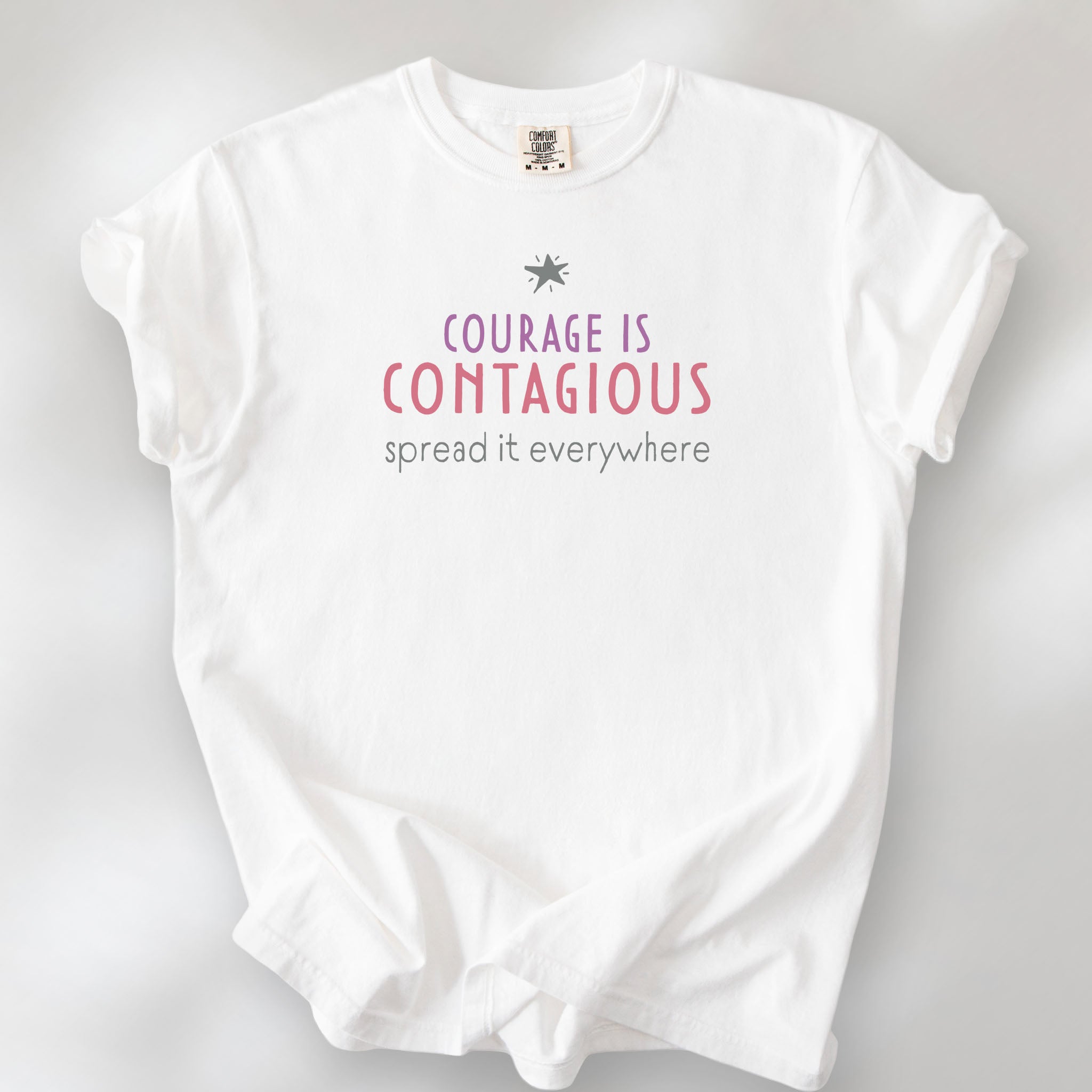 White t-shirt with the words “Courage Is Contagious – Spread It Everywhere” printed in soft purple, red, and gray.
