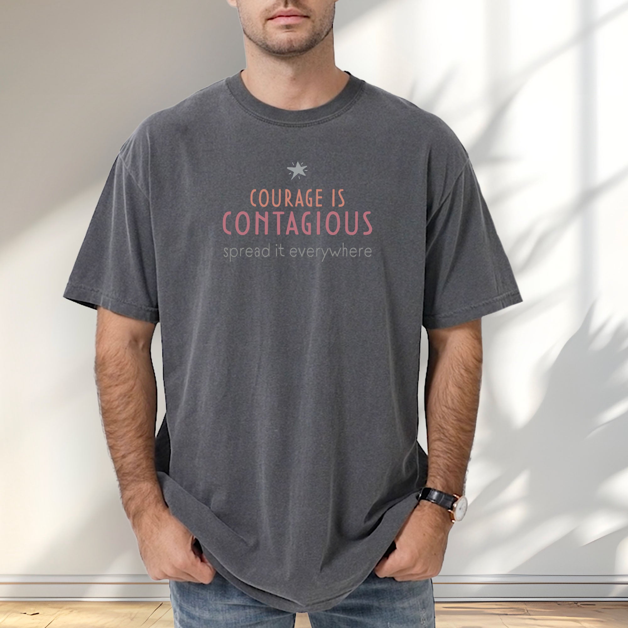Male model wearing pepper t-shirt with the words “Courage Is Contagious – Spread It Everywhere” printed in soft pink, coral, and gray.