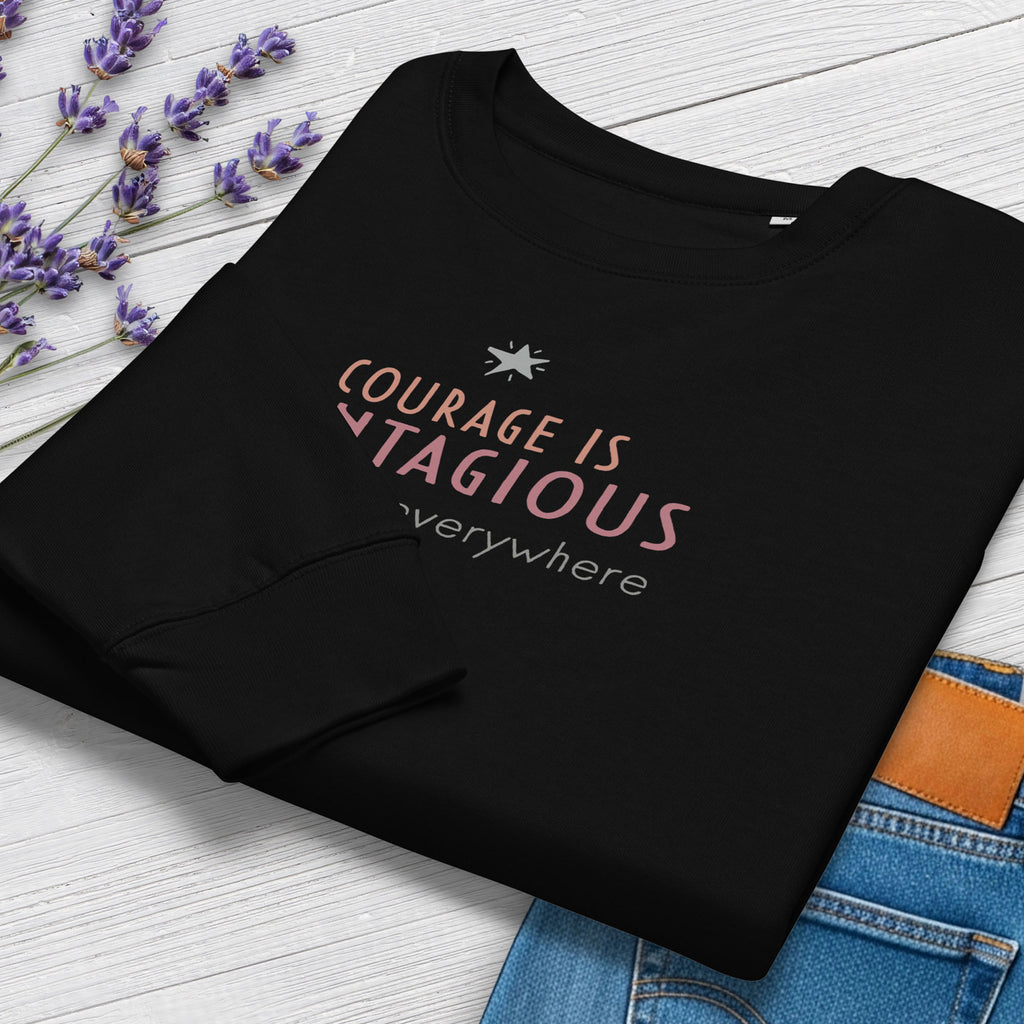 Folded black women's crewneck sweatshirt with minimalist stylized text that reads "courage is contagious – spread it everywhere". Closeup of cuffs and graphic, styled with jeans and flowers.