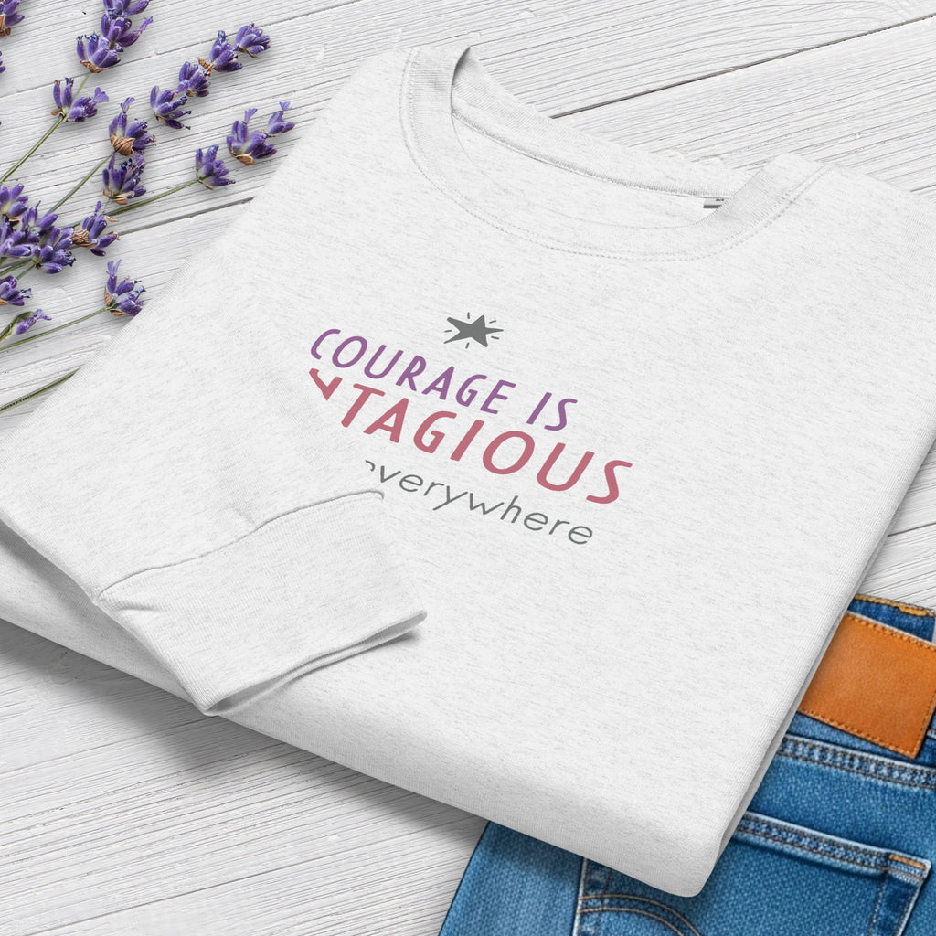 Folded grey women's crewneck sweatshirt with minimalist stylized text that reads "courage is contagious – spread it everywhere". Closeup of cuffs and graphic, styled with jeans and flowers.