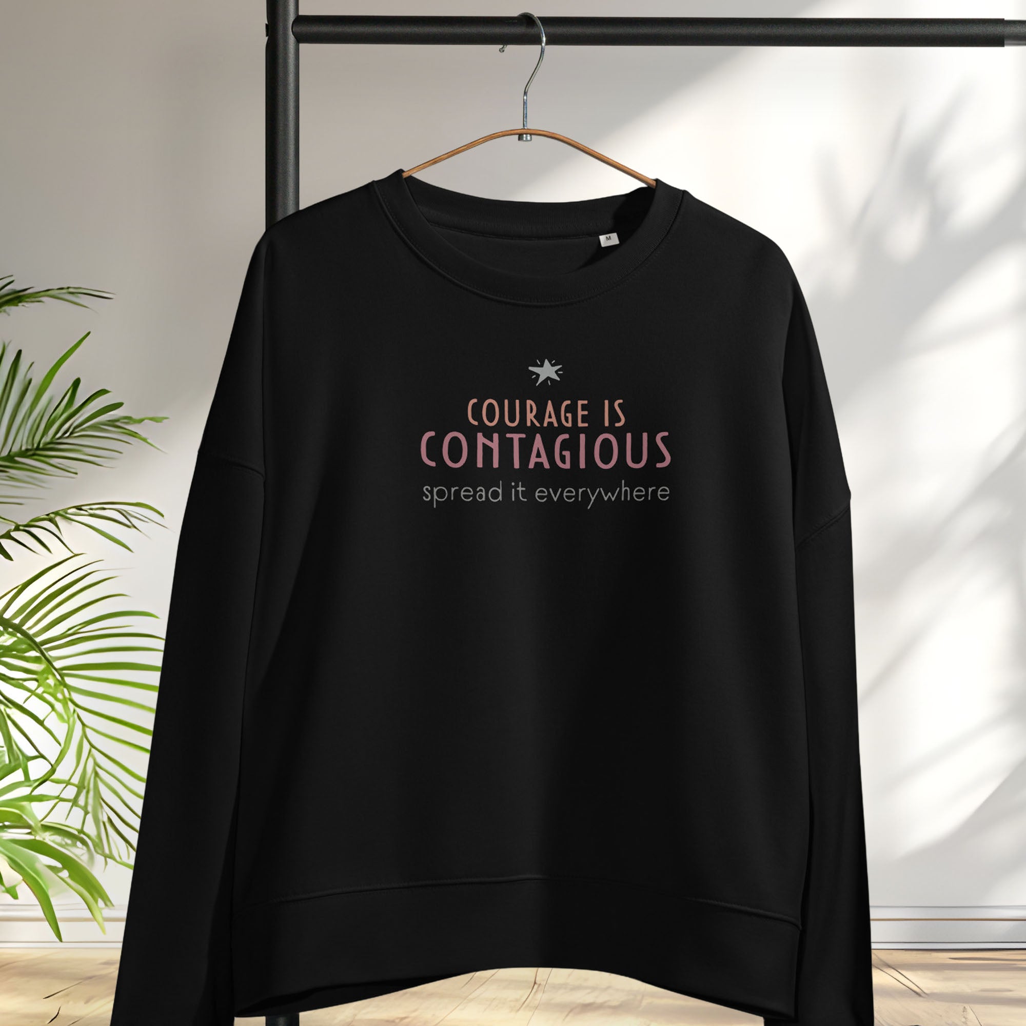 Black women's crewneck sweatshirt with minimalist stylized text that reads "courage is contagious – spread it everywhere". Hanging on a hanger.