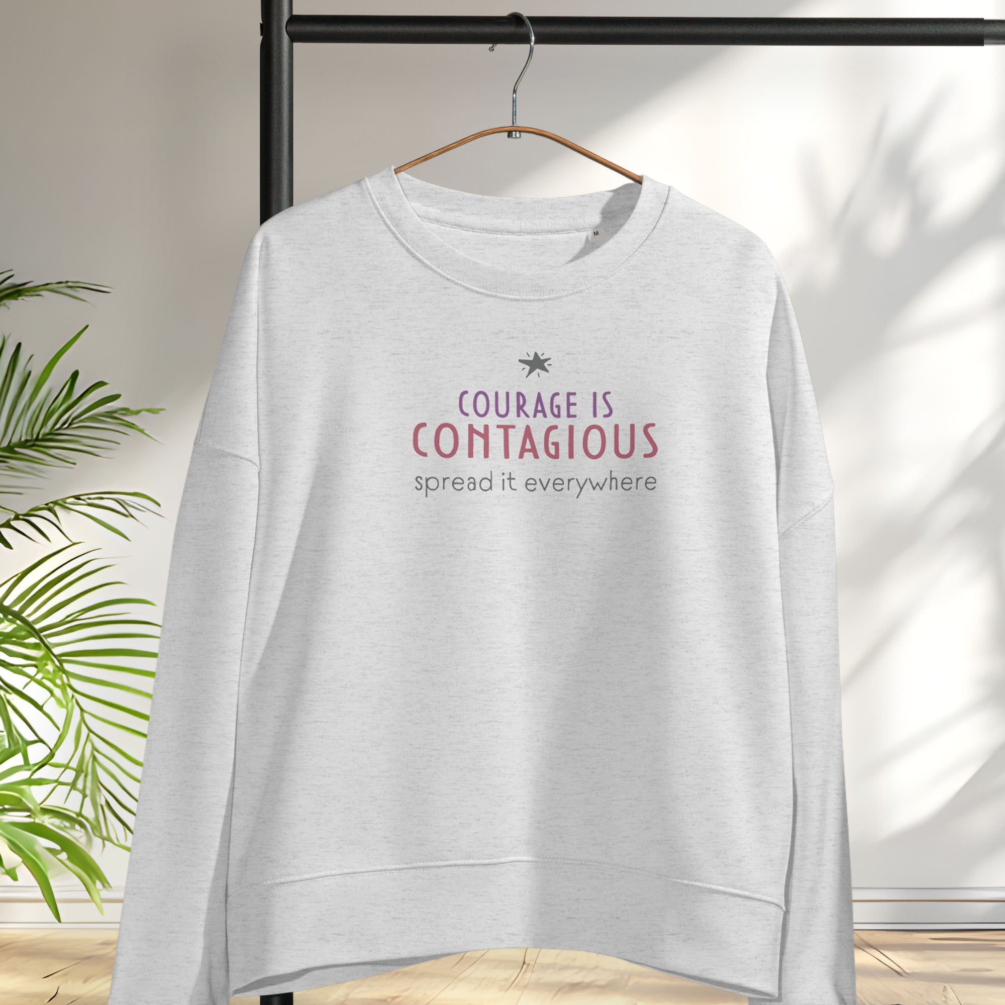 Grey women's crewneck sweatshirt with minimalist stylized text that reads "courage is contagious – spread it everywhere". Hanging on a hanger.