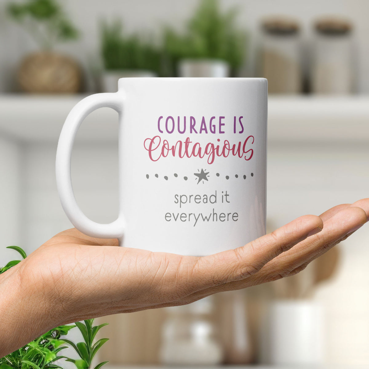 Mug with styled text graphic that reads "courage is contagious – spread it everywhere", held in the palm of a hand.
