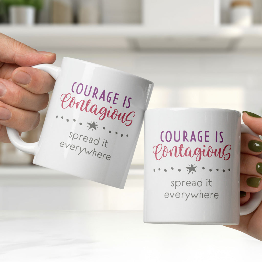 Two hands holding a white ceramic mug with styled text graphic that reads "courage is contagious – spread it everywhere", in a bright white kitchen.