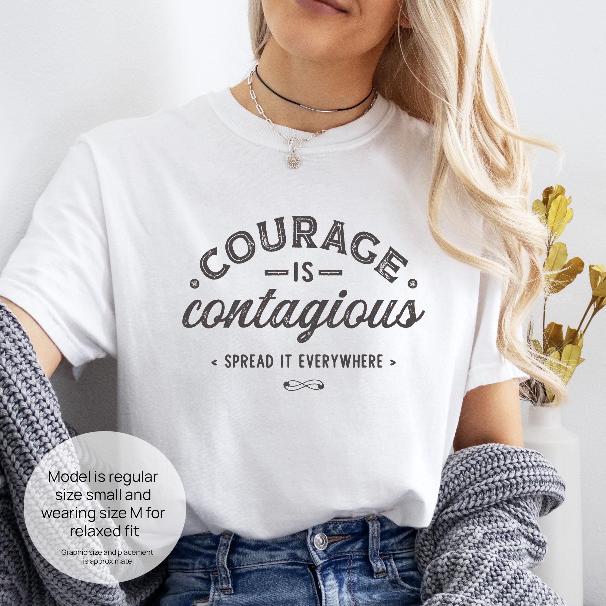 Female model wearing white "Courage Is Contagious Spread It Everywhere" t-shirt. Includes size and fit information for the model who is regular size small, and wearing a size M shirt for a relaxed fit.

